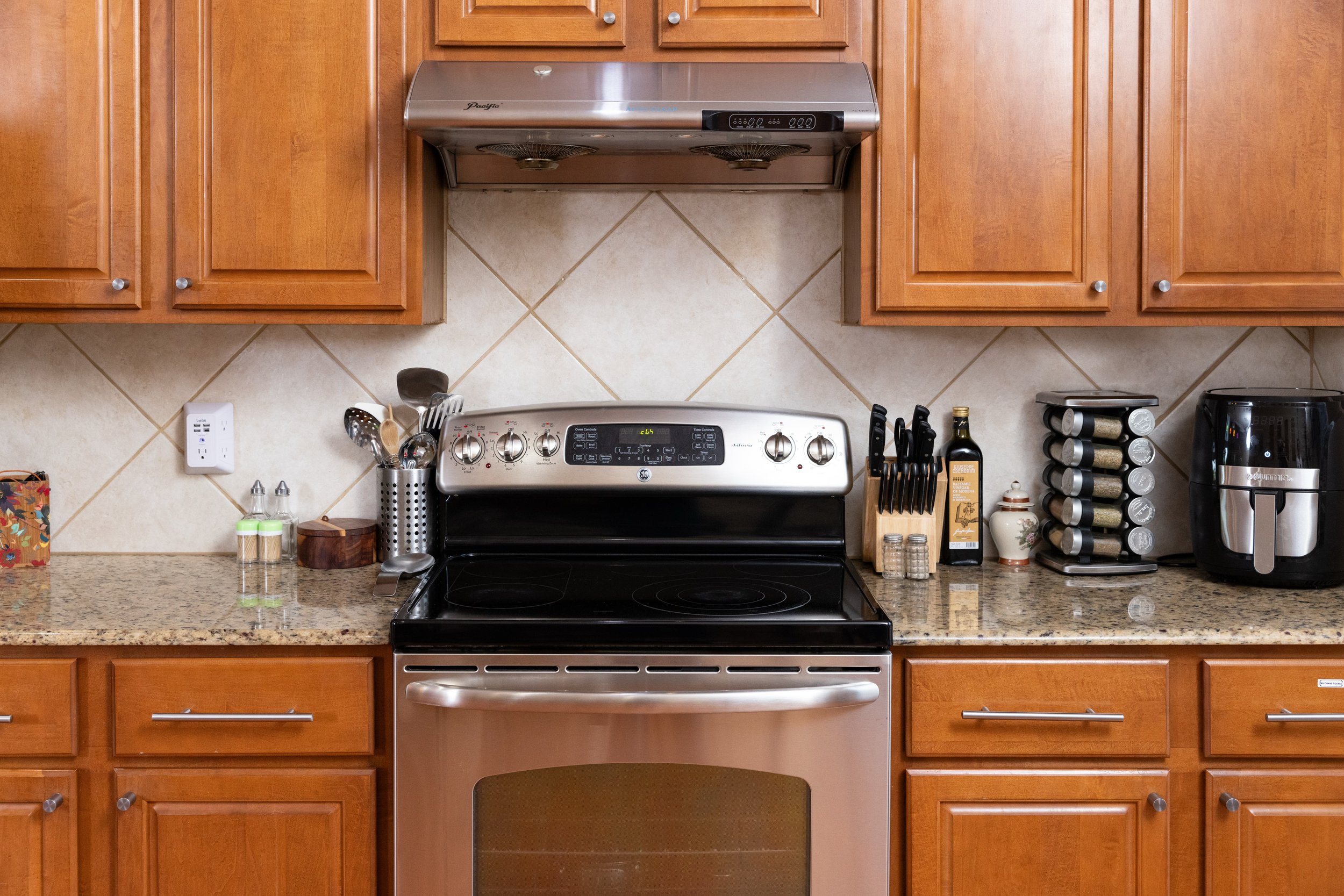 Modern kitchen with wooden cabinets, stainless steel stove, and granite countertops. Items include a knife block, spice rack, air fryer, cooking utensils, and olive oil bottle.