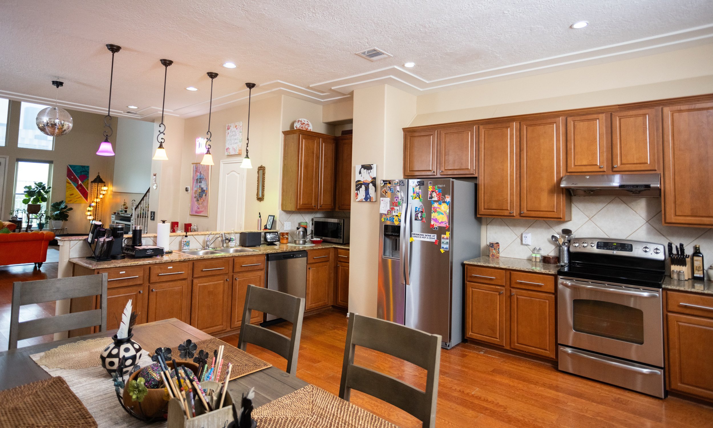 Modern kitchen with wooden cabinets, stainless steel appliances, and colorful pendant lights. Dining table with decorative items in foreground.