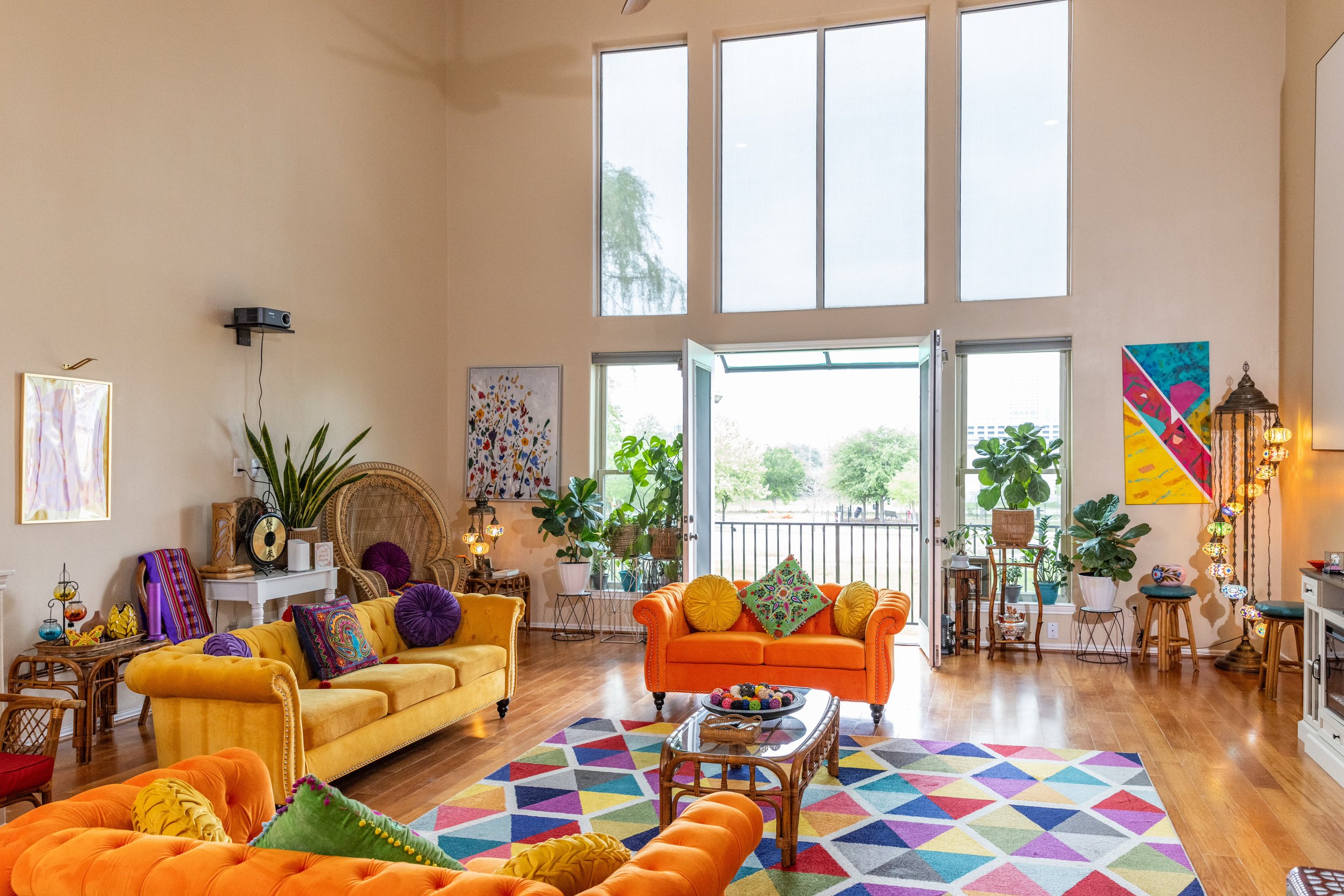 Brightly colored living room with large windows, featuring orange and yellow sofas, a colorful geometric rug, and abundant potted plants. The room includes vibrant artwork, a glass coffee table, and a mix of eclectic decor items.