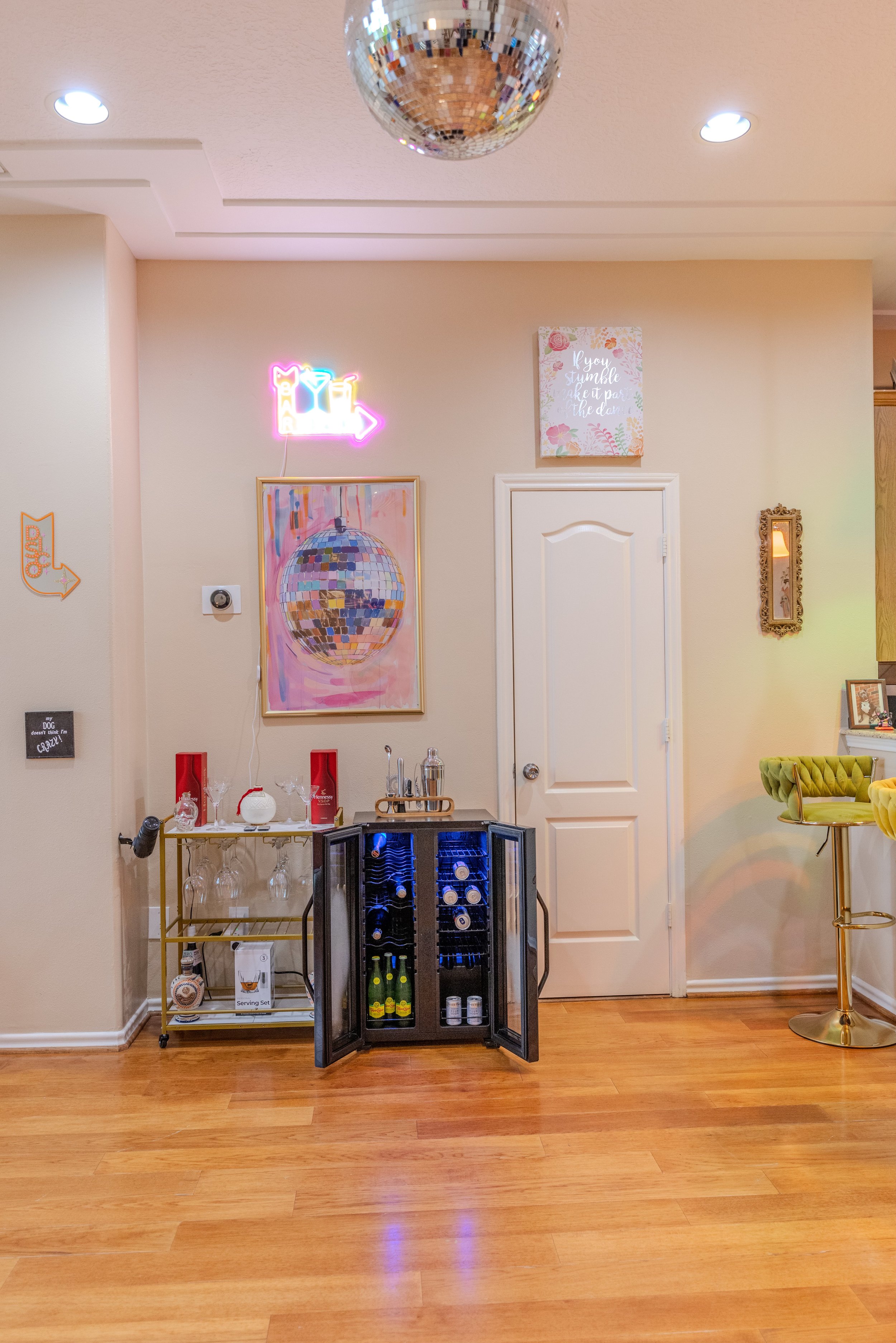 A home bar setup featuring a minibar with open doors displaying bottles, a glassware cart, wall art including a neon sign, a mirrored disco ball, and a plush bar stool. The room has wooden flooring and neutral walls.