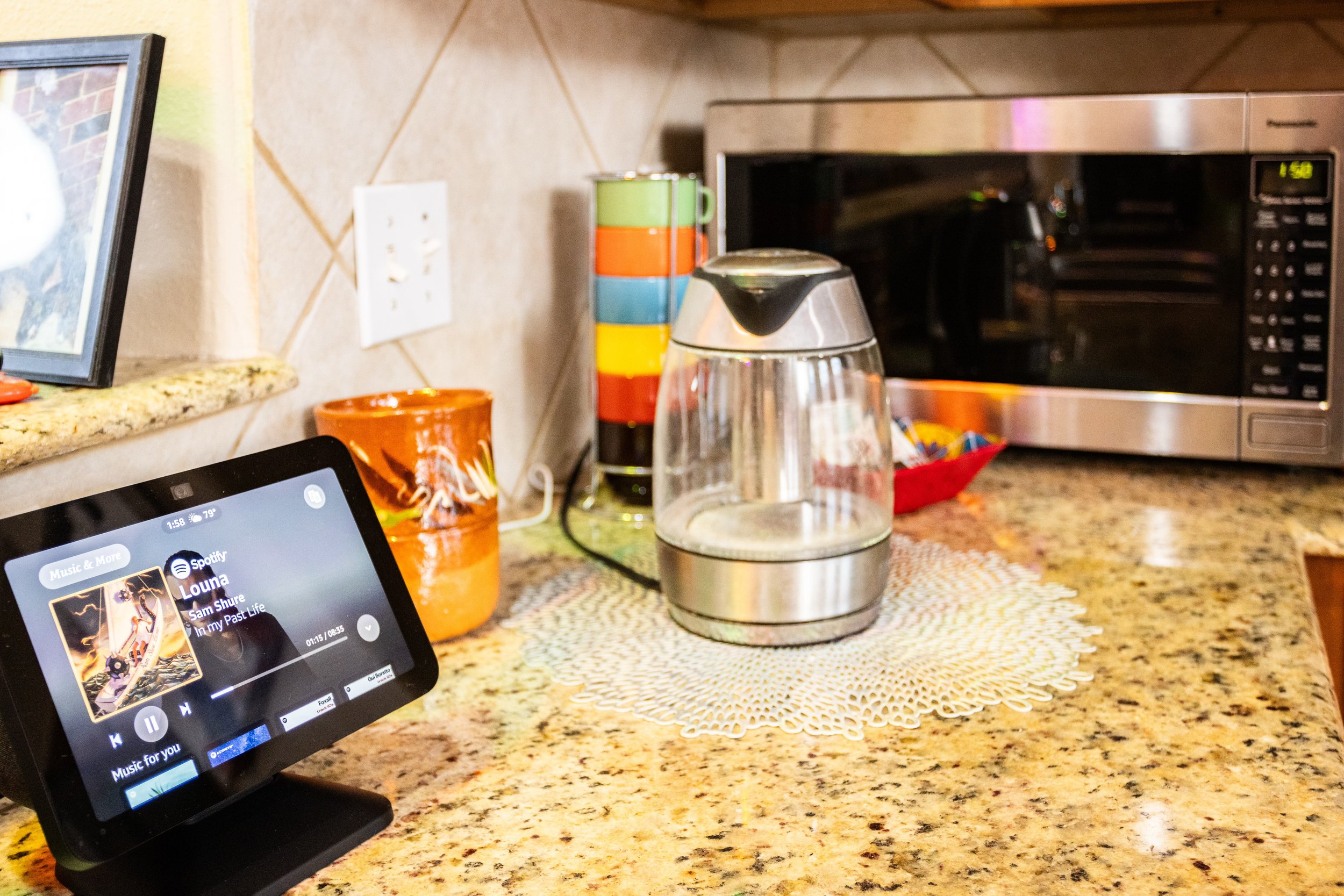 Kitchen countertop with a smart display showing Spotify, an electric kettle, colorful stacked mugs, a microwave, and a photo frame.