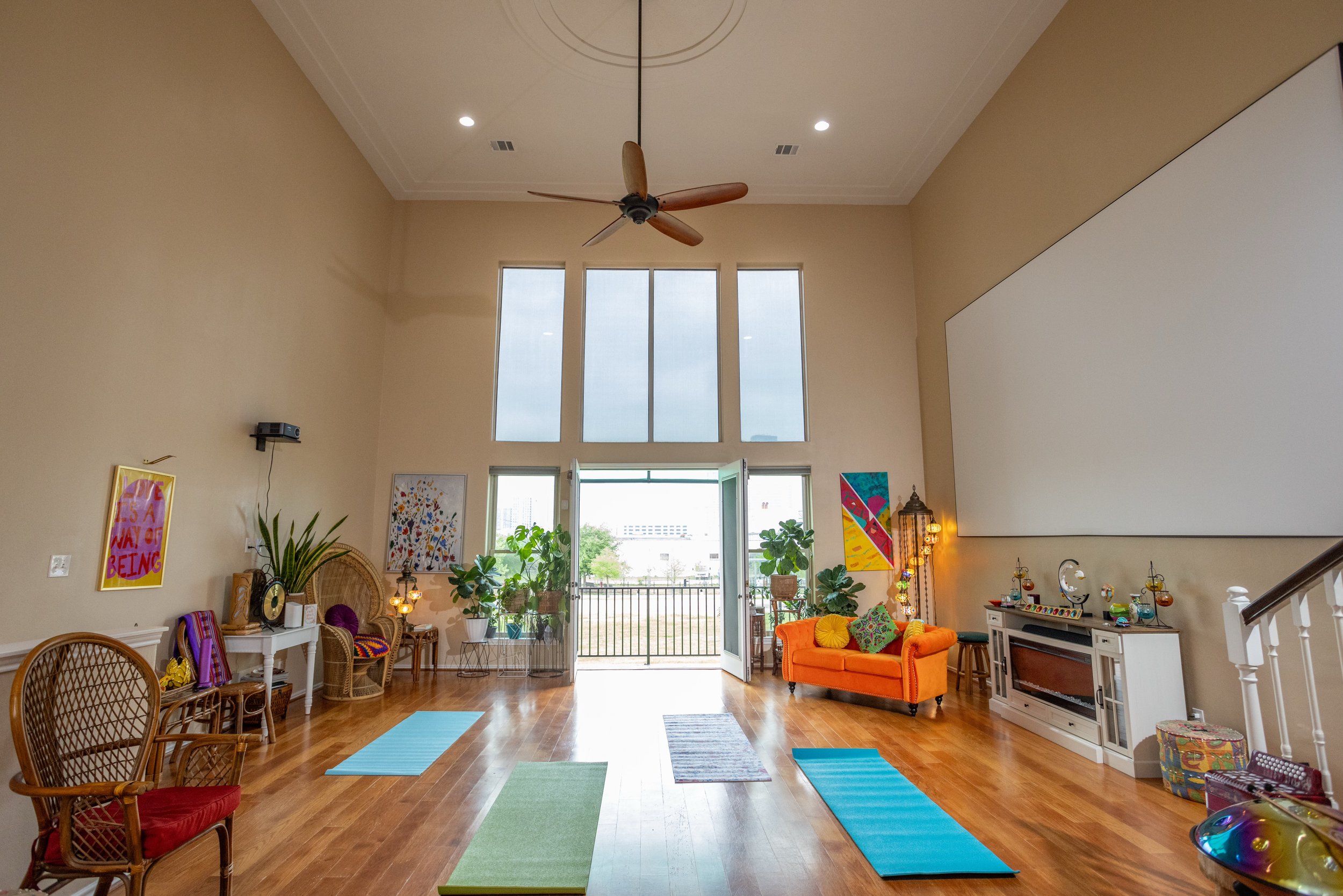 Spacious room with high ceilings, an orange sofa, colorful yoga mats on wooden floor, large windows, plants, and artwork. A ceiling fan hangs above, and there's a decorative fireplace against one wall.