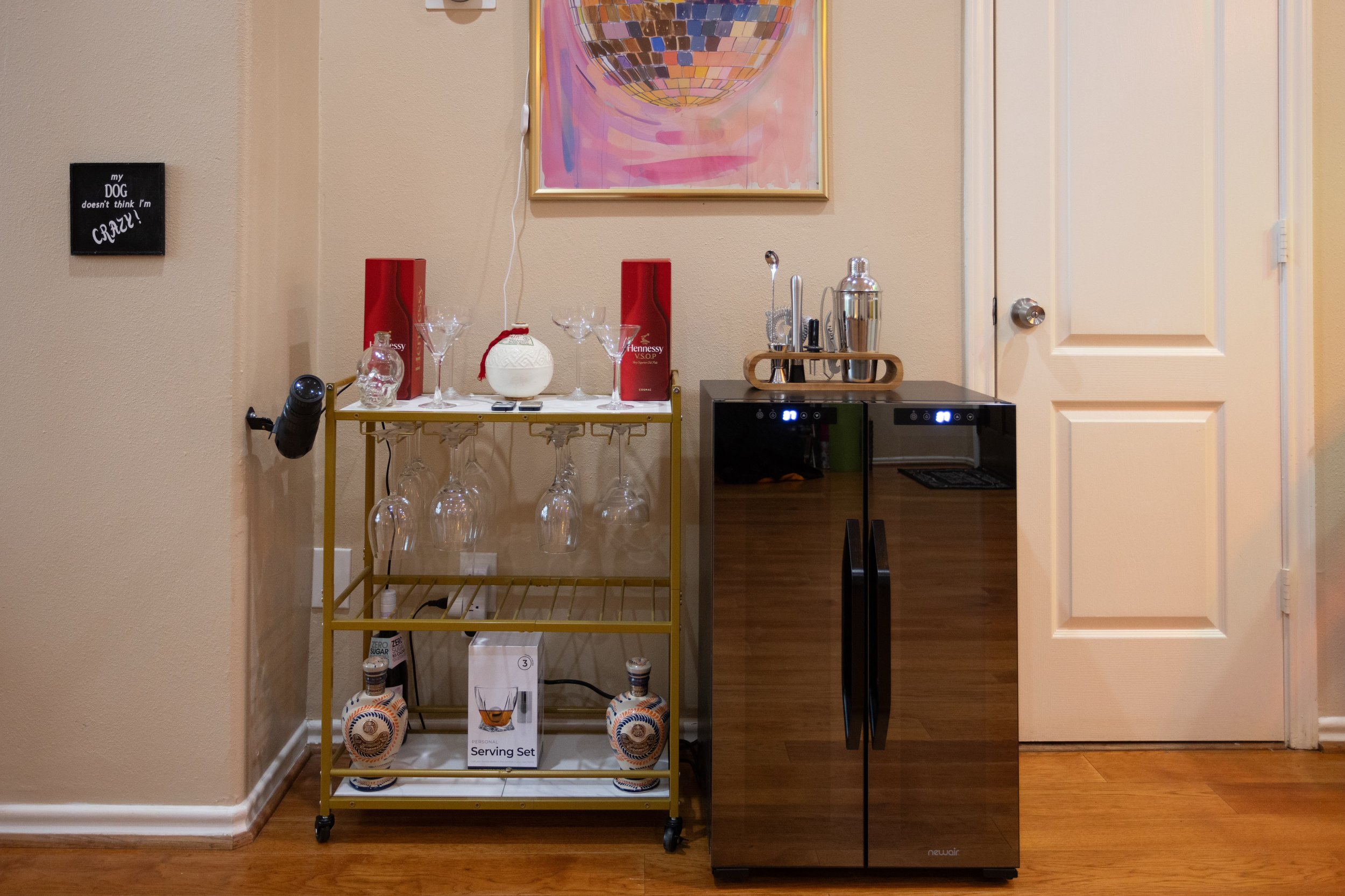 Home bar setup with a cart holding wine glasses, liquor bottles, and a serving set next to a double-door wine refrigerator. Above is a colorful abstract painting, and a humorous sign is on the wall.