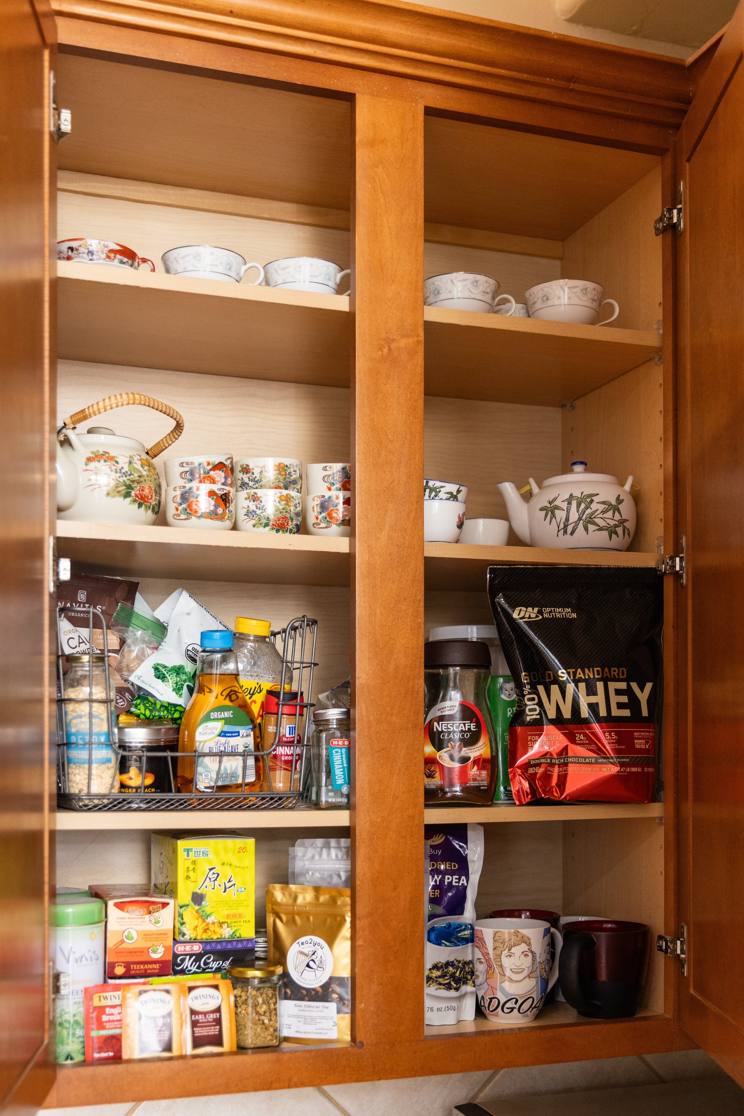 Interior of a wooden kitchen cabinet with assorted tea cups, teapots, packaged food items, coffee containers, whey protein powder, and jars on shelves.