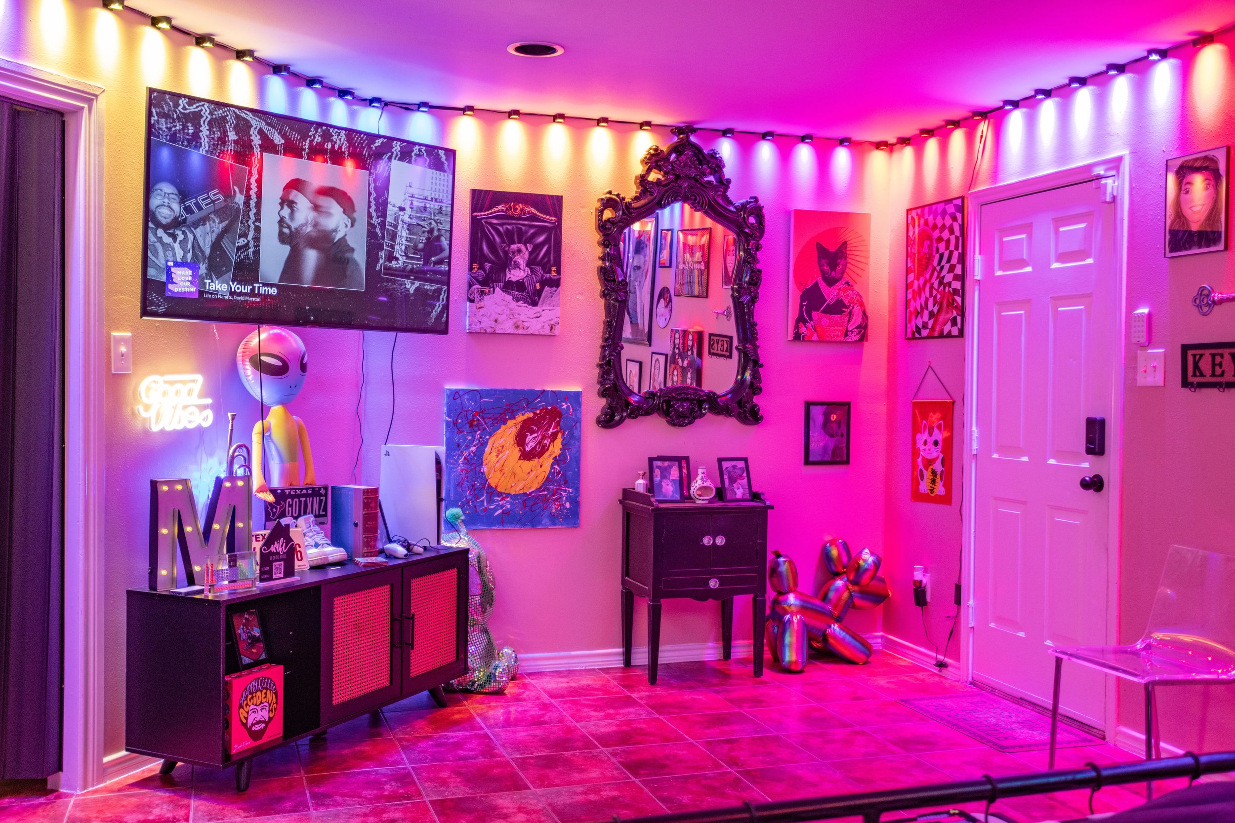 A brightly lit room with colorful LED lights, featuring a wall-mounted TV displaying album artwork, a cabinet with decor including a glowing 'Good Vibes' sign and an alien figure, assorted wall art, and a baroque mirror. A small table with framed pho