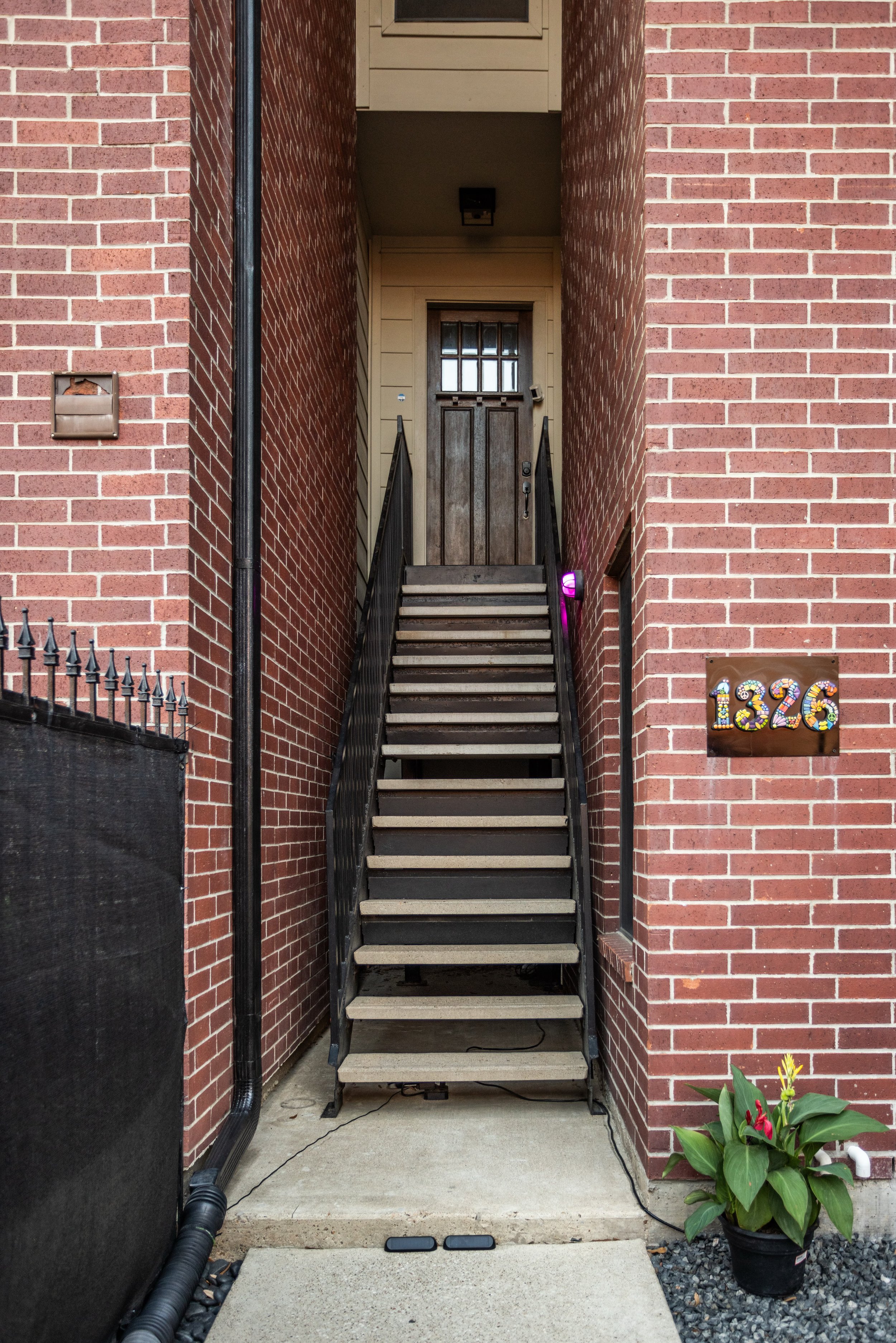 Brick exterior of a building with numbered address plaque "1326" next to a staircase leading to a wooden door. A potted plant is placed near the entrance, and a narrow iron railing lines the steps.