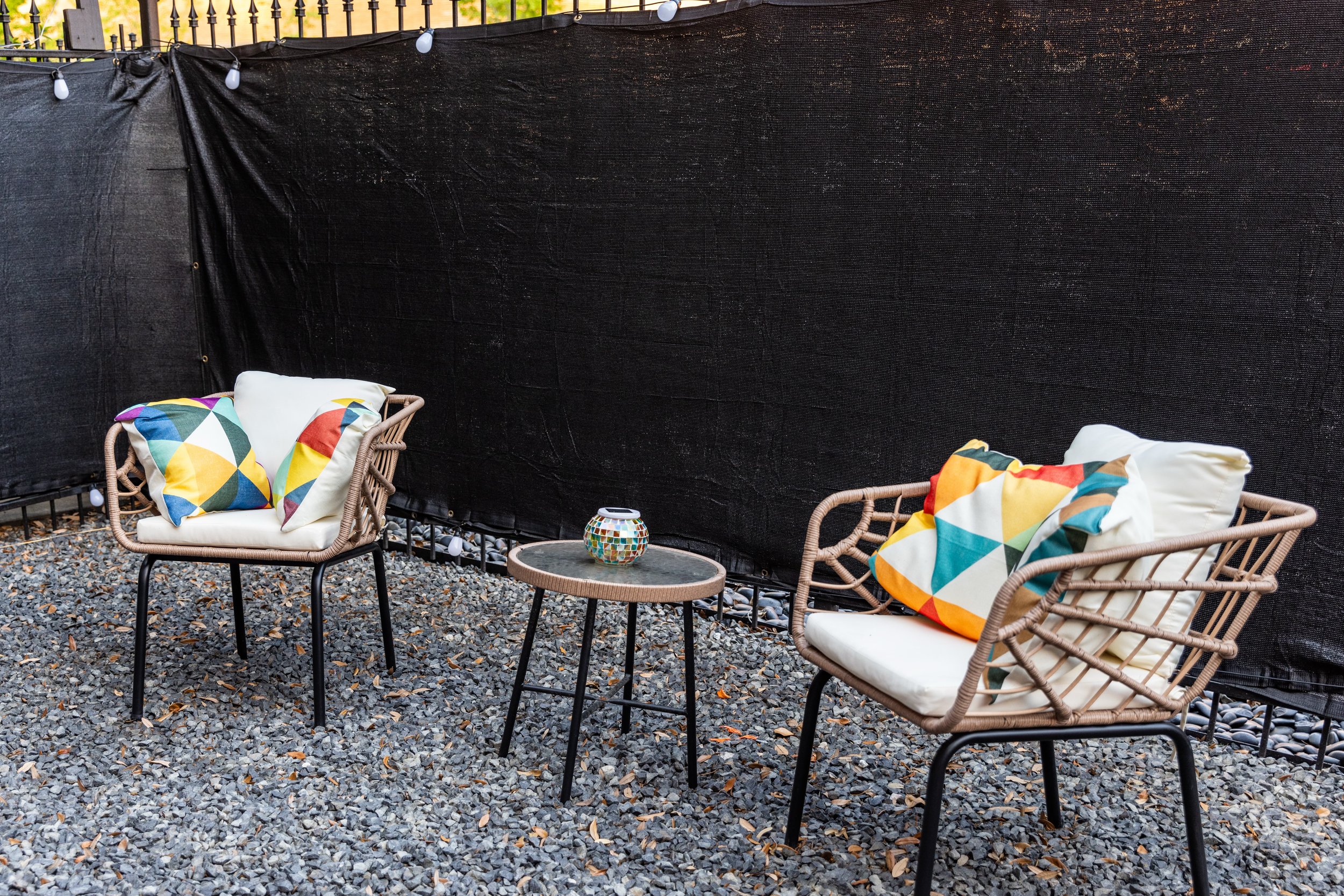 Outdoor seating area with two wicker chairs featuring colorful geometric cushions, separated by a small round table with a mosaic-patterned candle holder, set on gravel in front of a black fabric fence.