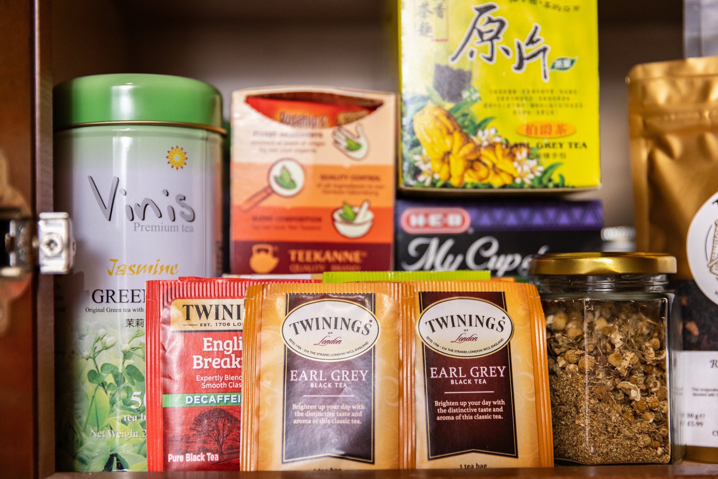 Variety of tea packages and jars in a cupboard, including Twinings English Breakfast and Earl Grey, Vinis Jasmine Green Tea, and a jar of loose tea.