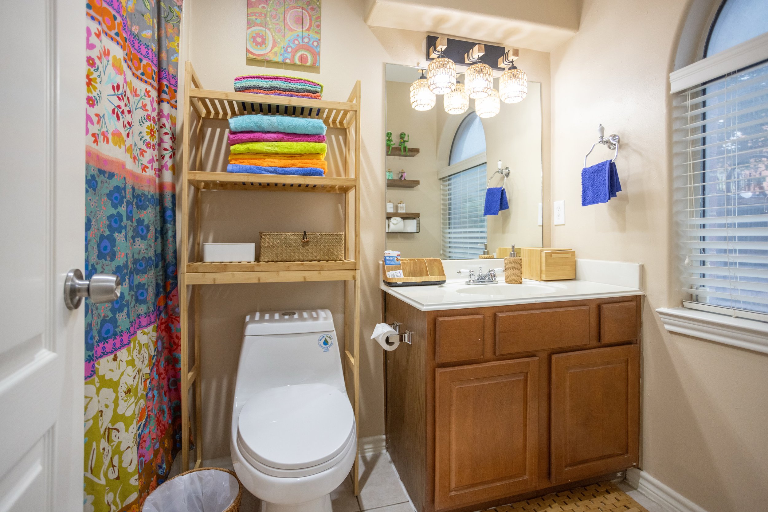 Bathroom interior with a toilet, wooden shelf holding colorful towels, wicker basket, single sink vanity with mirror, decorative lights, floral shower curtain, and blue towel on a wall hook.