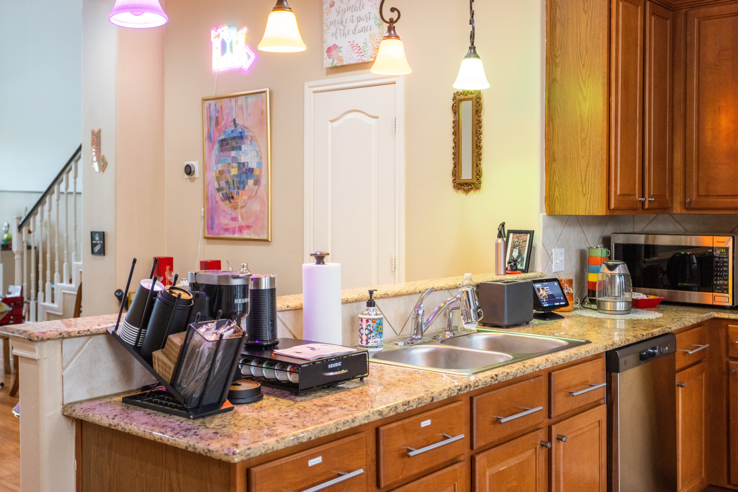 Modern kitchen counter with coffee maker, paper towel holder, dishwasher, microwave, sink, decorative lighting, colorful artwork, and wooden cabinets.