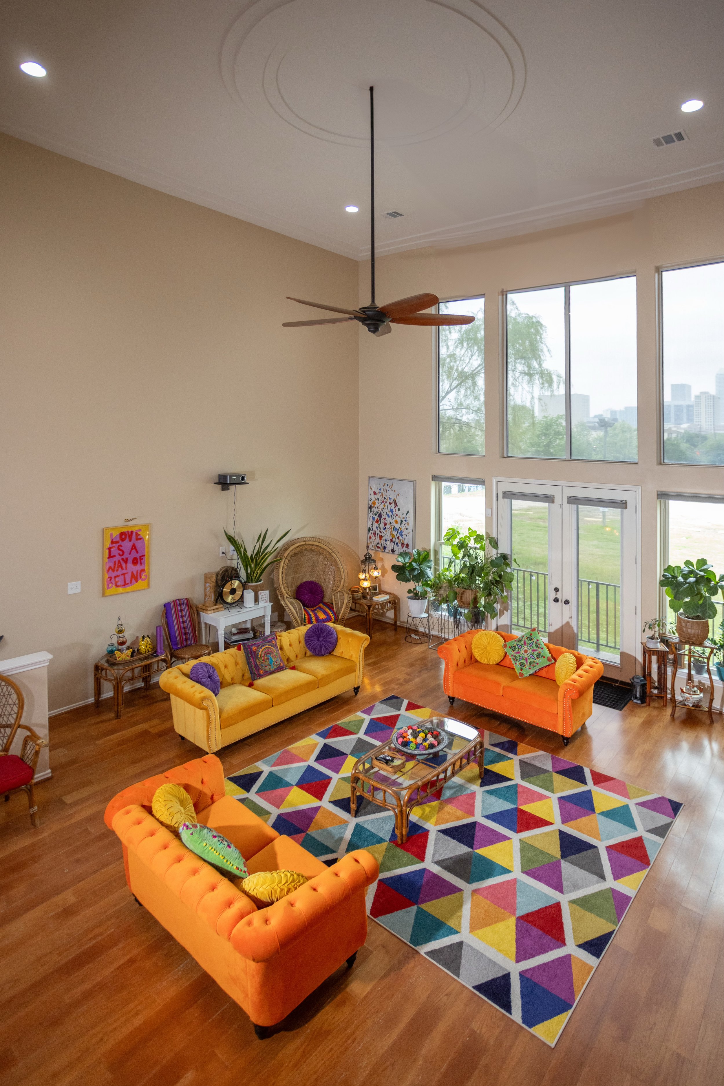Modern living room with colorful furniture, including yellow and orange sofas, a geometric patterned rug, and large windows. Plants and colorful decor enhance the space.