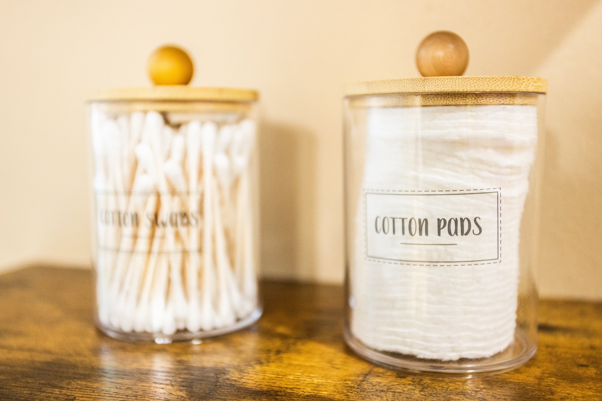 Glass jars with wooden lids on a table, one labeled 'Cotton Swabs' and the other 'Cotton Pads.'