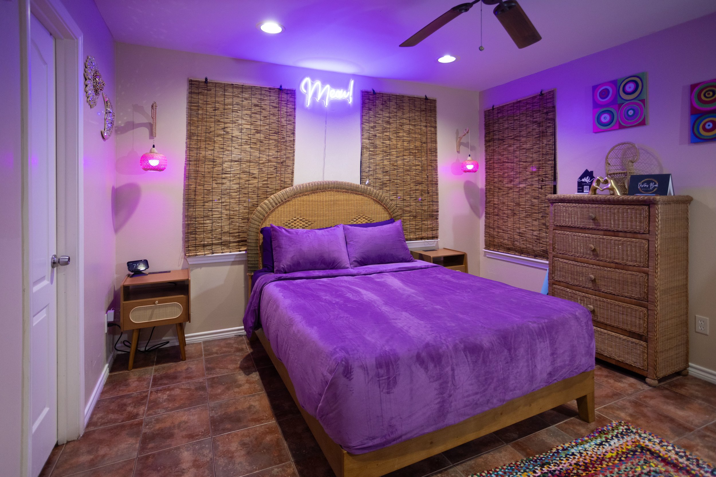 Cozy bedroom with purple bedding, neon "Meow" sign, wicker headboard and dresser, woven blinds, wooden nightstands, pink pendant lights, colorful wall art, and tiled floor.