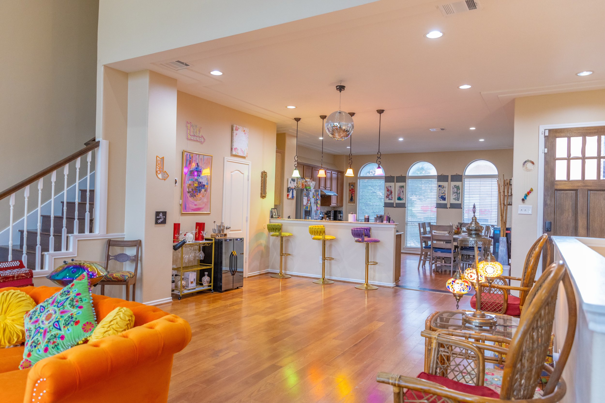Colorful, retro-themed open-plan living room and kitchen with orange sofa, multicolored bar stools, pendant lights, disco ball, and decorative pillows.