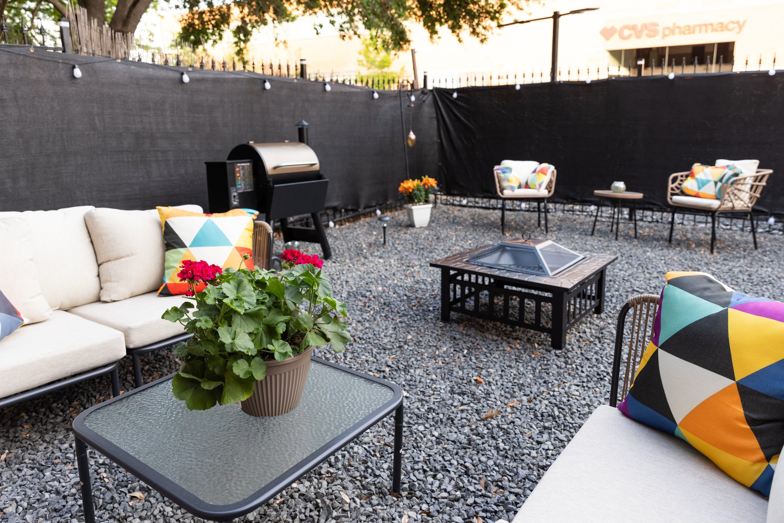 Outdoor patio with seating, colorful geometric cushions, fire pit, grill, potted plants, and decorative lights on gravel ground.