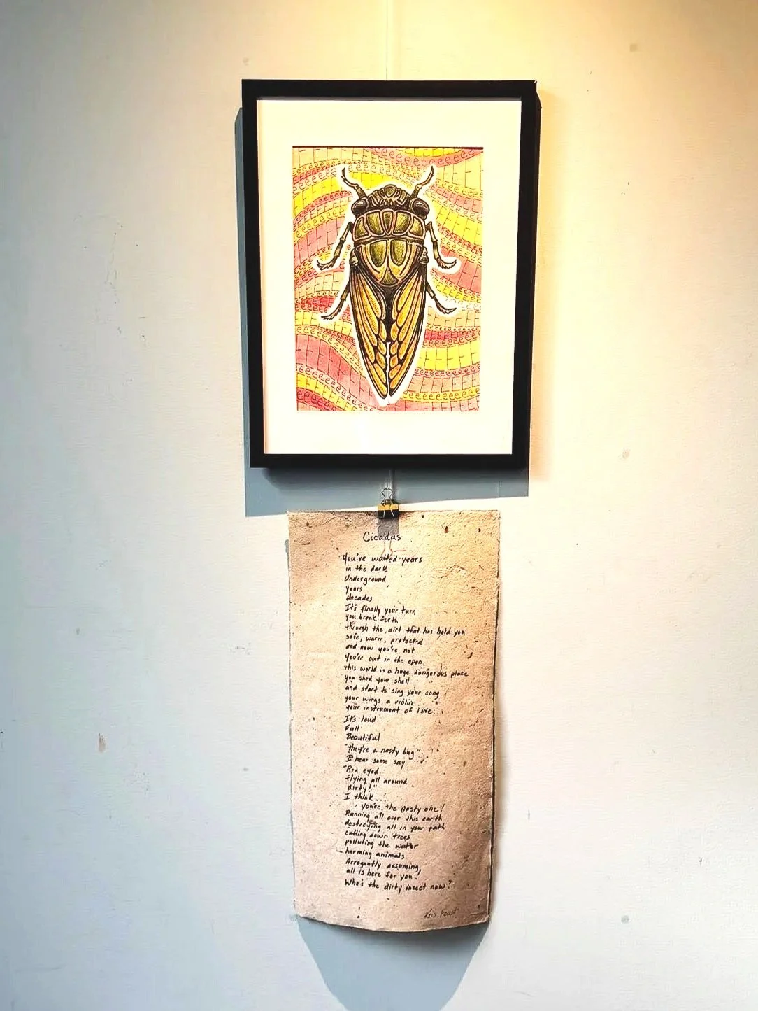 Colorful framed artwork of a beetle with yellow and pink background, hanging on a white wall, with a handwritten poem on brown paper hanging below it.