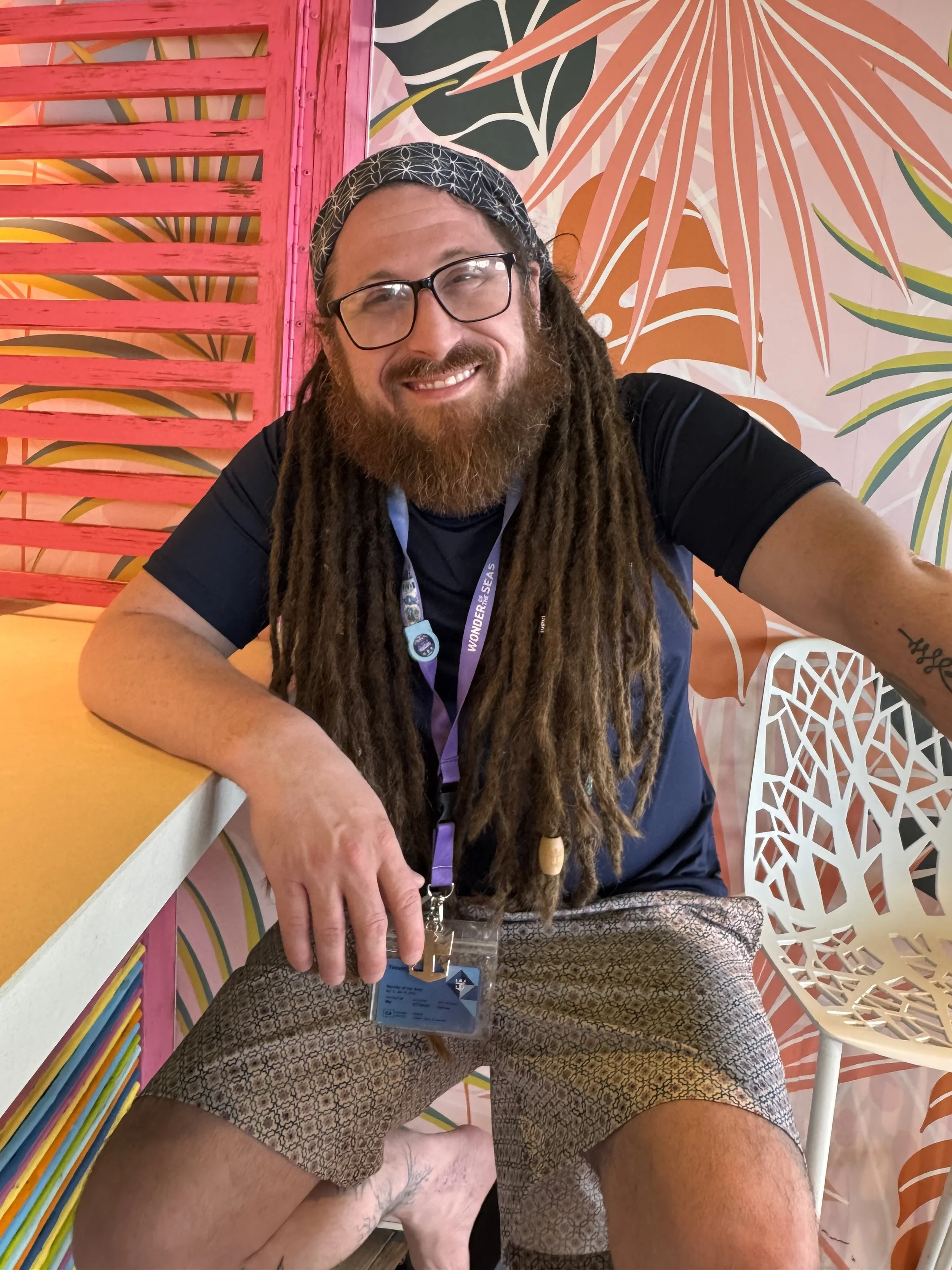 Chef Tim with long dreadlocks, glasses, and a beard smiling, sitting in front of colorful, tropical-themed wall art.