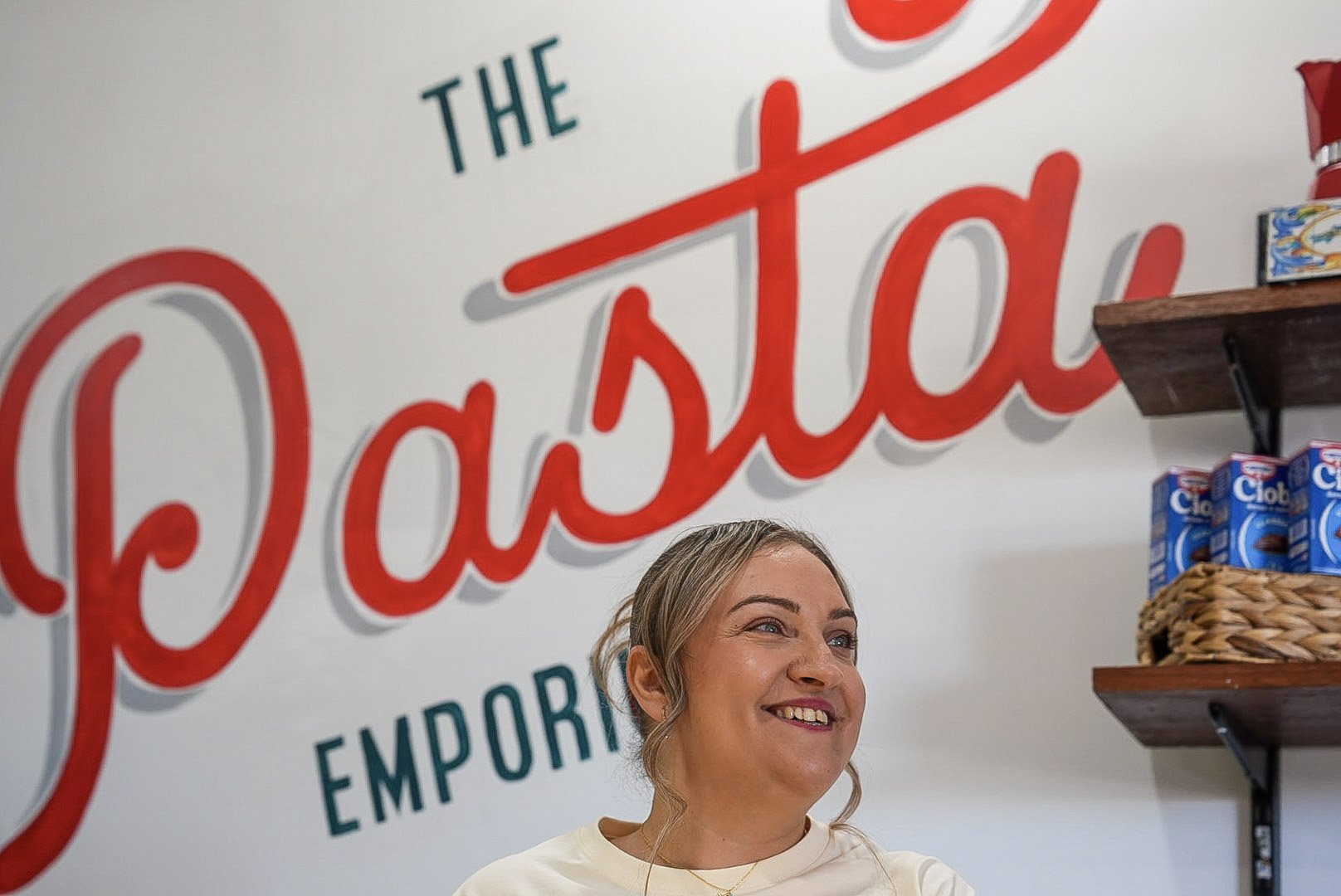 Smiling woman in front of a wall sign that says 'The Dabbler Emporium' with shelves of cereal boxes behind her.