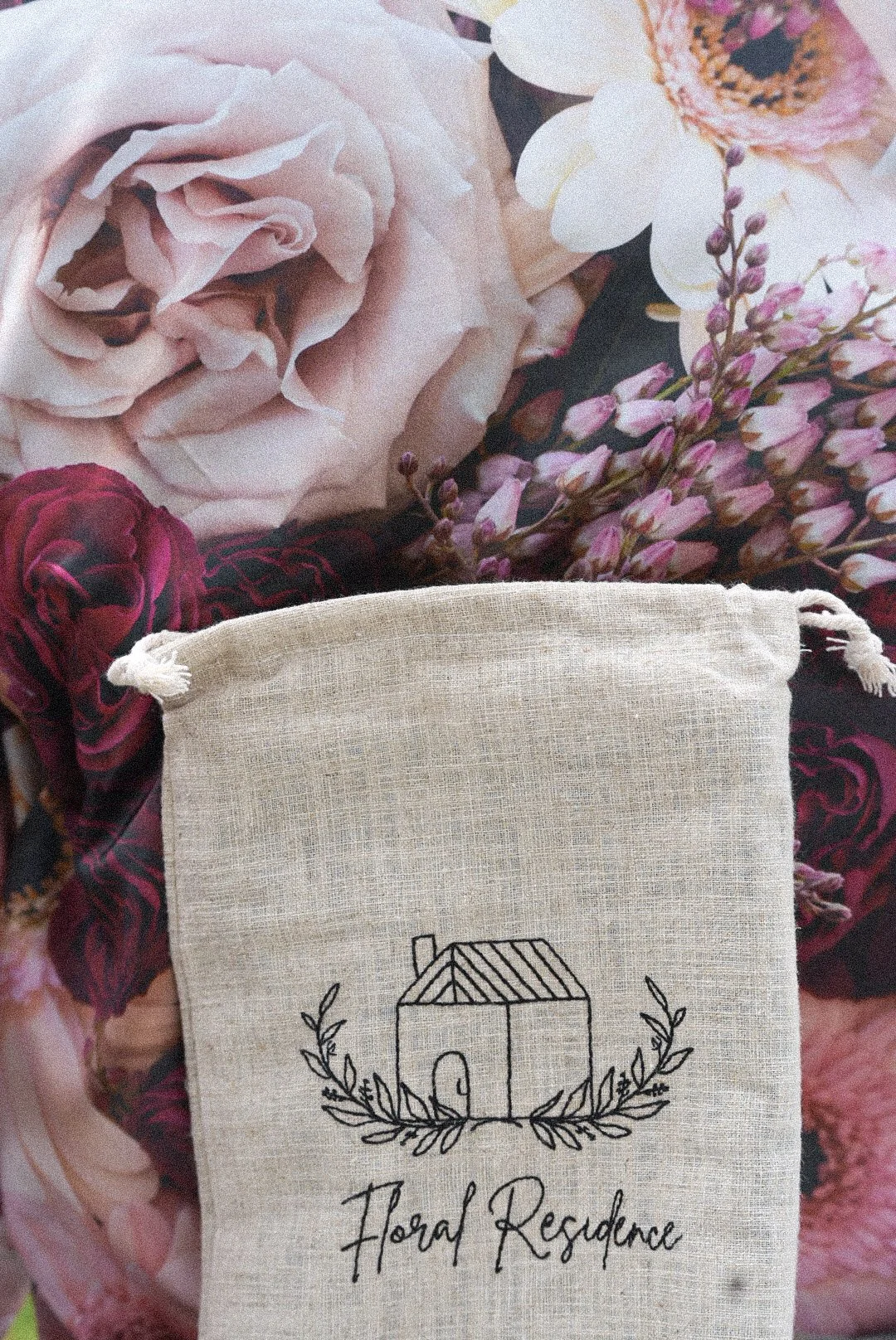 A beige linen bag with a black embroidered house and leaves, placed among a variety of pink and white flowers.