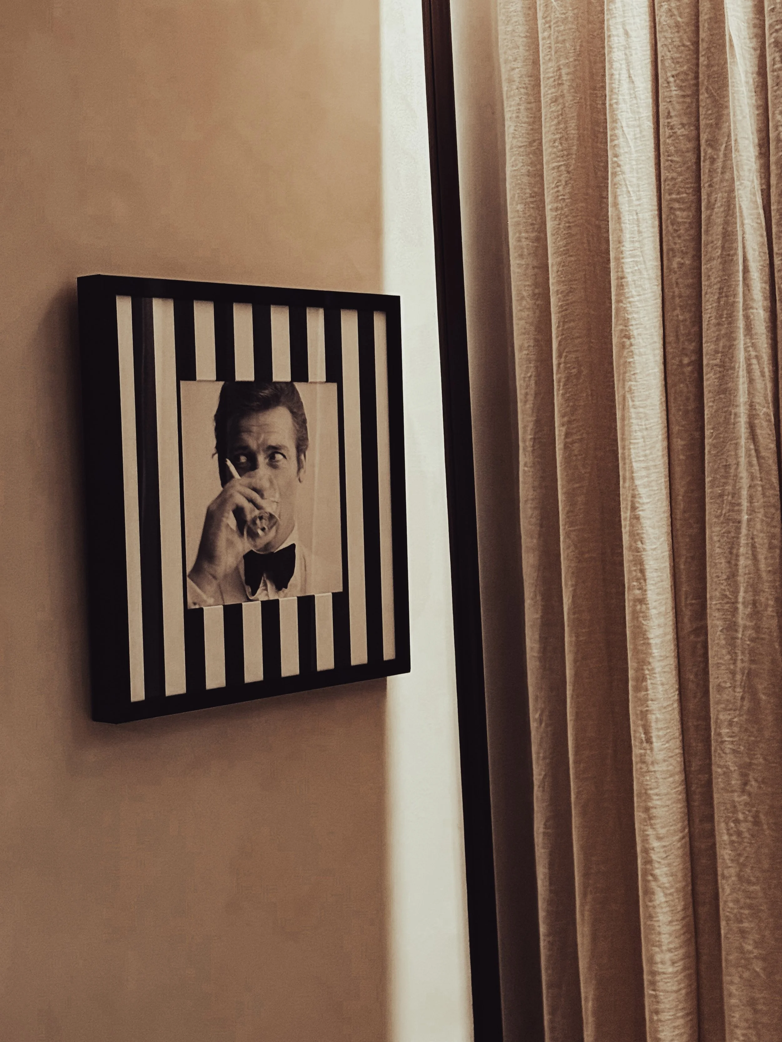 A framed black and white photo of a man in a tuxedo drinking from a glass, with black and white vertical stripes in the background, hanging on a beige wall next to light-colored curtains.