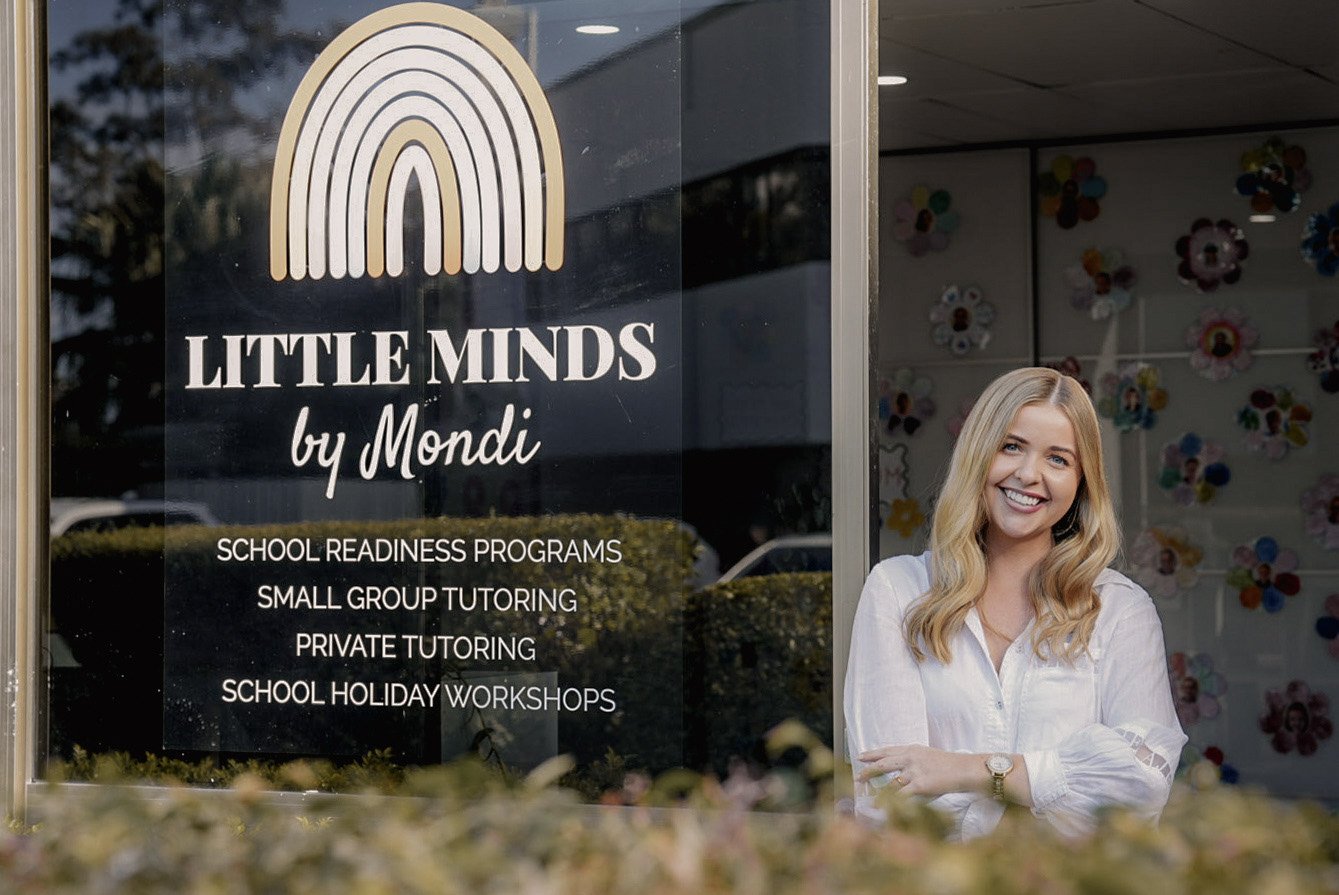 Sign for Little Minds by Mondi offering school readiness programs, small group tutoring, private tutoring, and school holiday workshops, with a woman smiling outside the business.