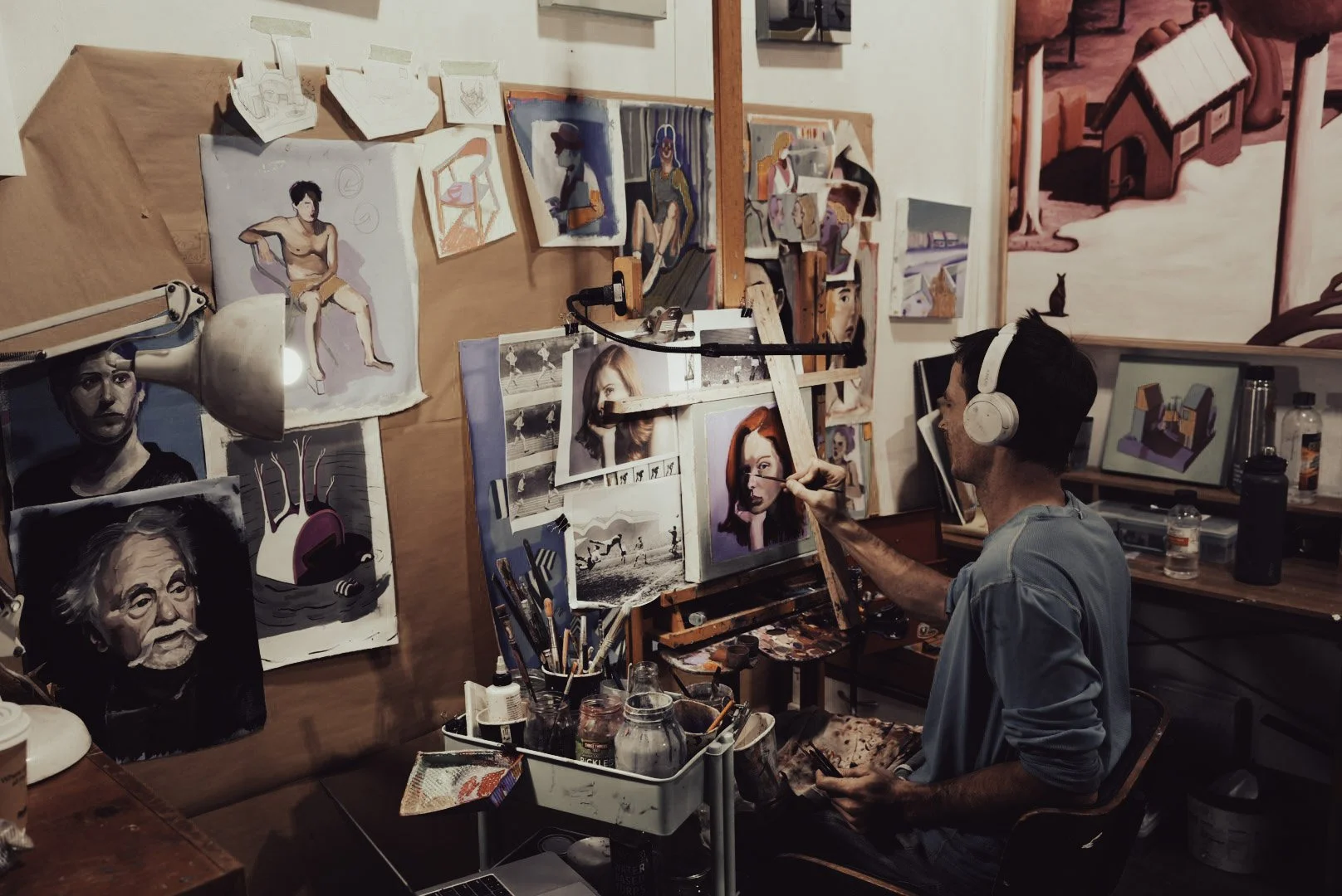 An artist wearing headphones is painting a portrait on a canvas in an art studio filled with various artwork, sketches, and art supplies.