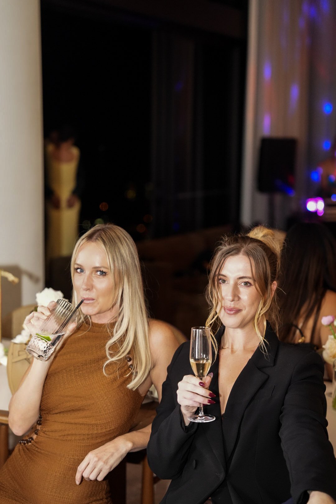 Two women at a social gathering, one drinking from a glass with a straw and the other holding a glass of champagne, in a dimly lit room with colorful lights.