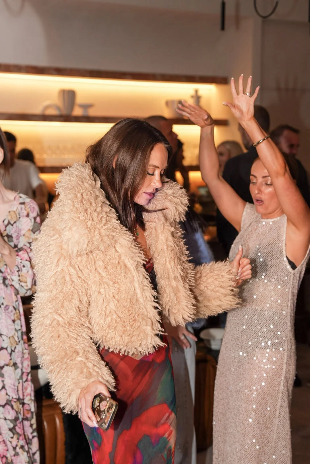 Women dancing at a social gathering or party in a warmly lit room, with shelves in the background.
