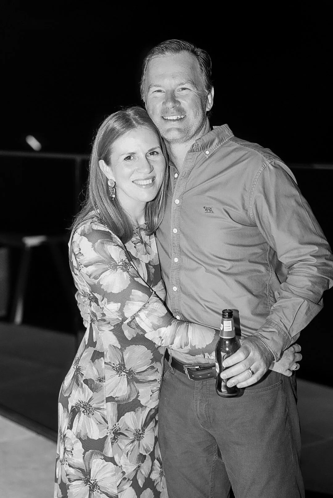 A smiling couple, a woman in a floral dress and a man in a button-up shirt, hugging and holding drinks at an outdoor evening event.