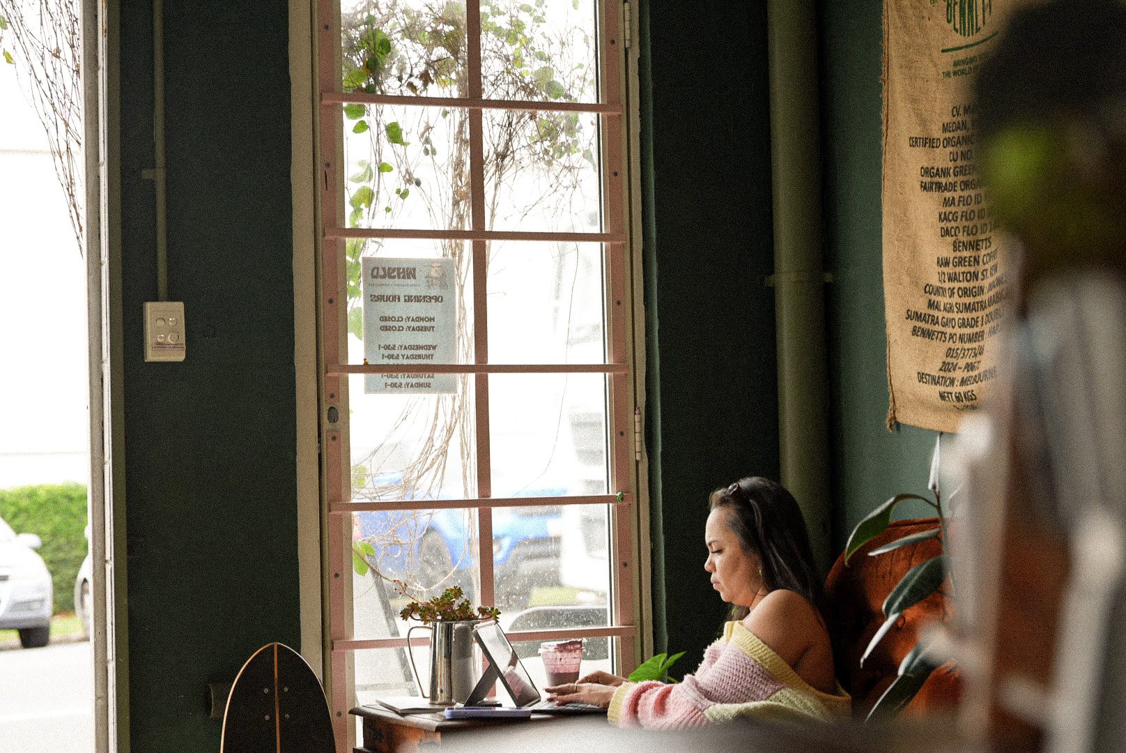 Woman working on a laptop inside a cafe or shop, sitting at a wooden table near a large window with pink blinds, with plants and a poster on the wall.