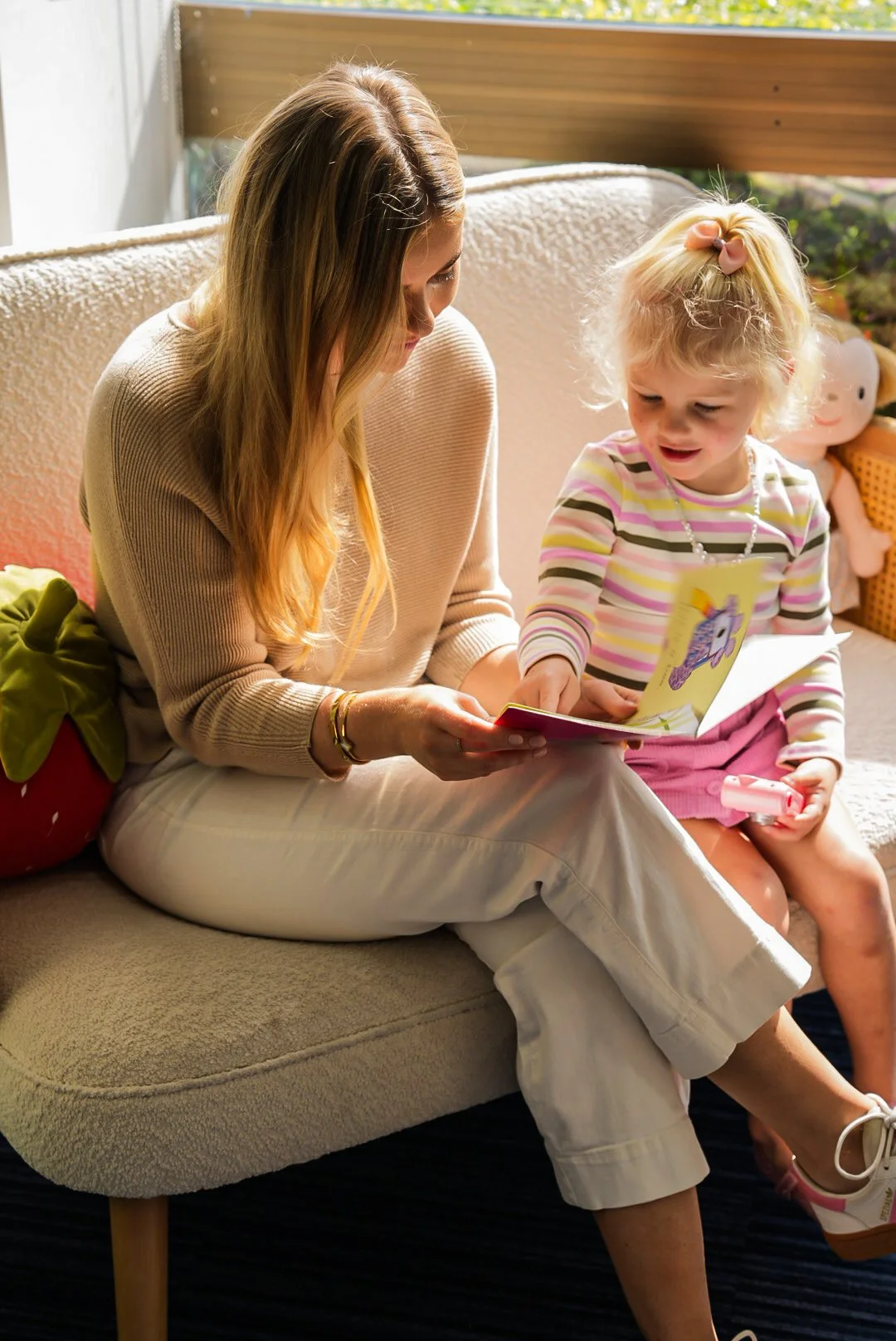 A woman and a young girl sitting on a beige couch inside a home, looking at a picture book together. The woman is wearing a light beige sweater and white pants, while the girl wears a striped long-sleeve shirt and a pink skirt with a pink bow in her 