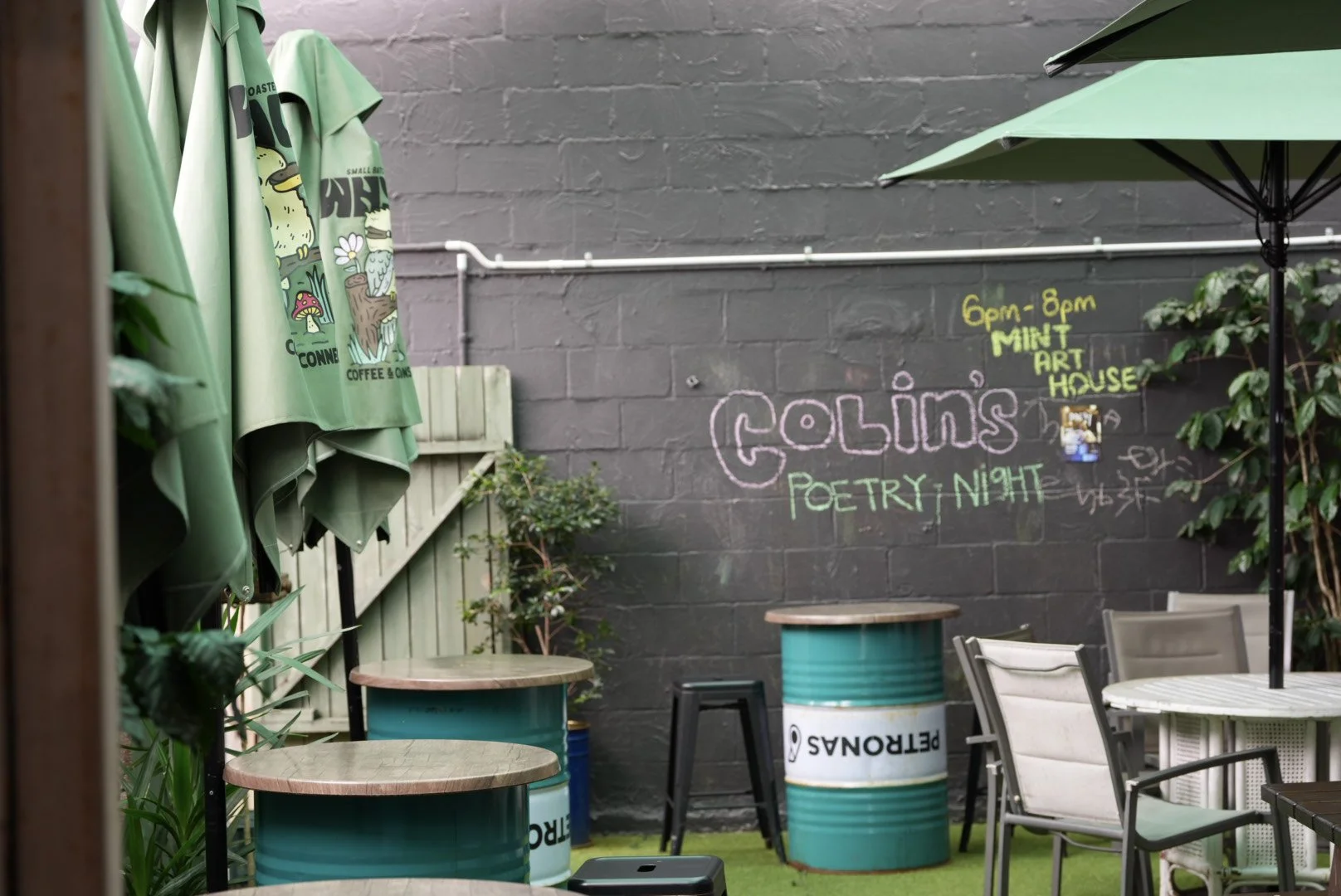 An outdoor seating area with green umbrellas, trees, and colorful painted barrels used as tables. A dark gray brick wall features neon-colored chalk writing promoting a poetry night event at the Mint Art House, with some chairs and tables arranged fo