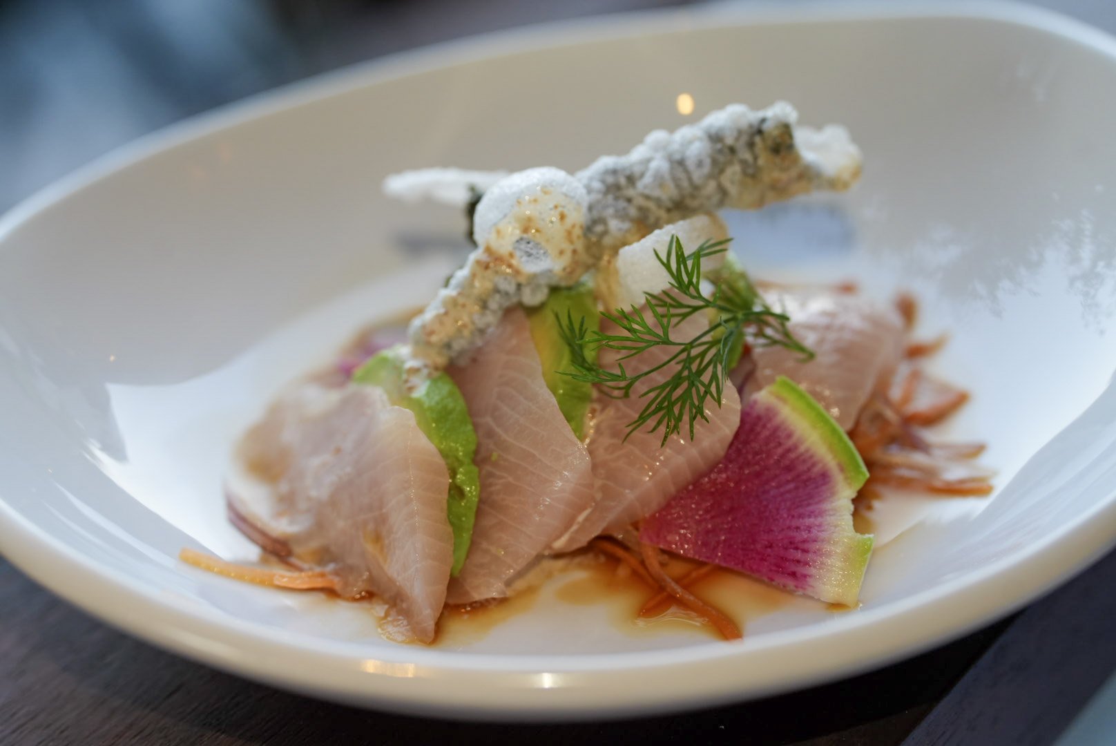 A gourmet dish featuring sliced raw fish garnished with fresh herbs, thinly sliced radish, and crispy fried garnish on a white plate.