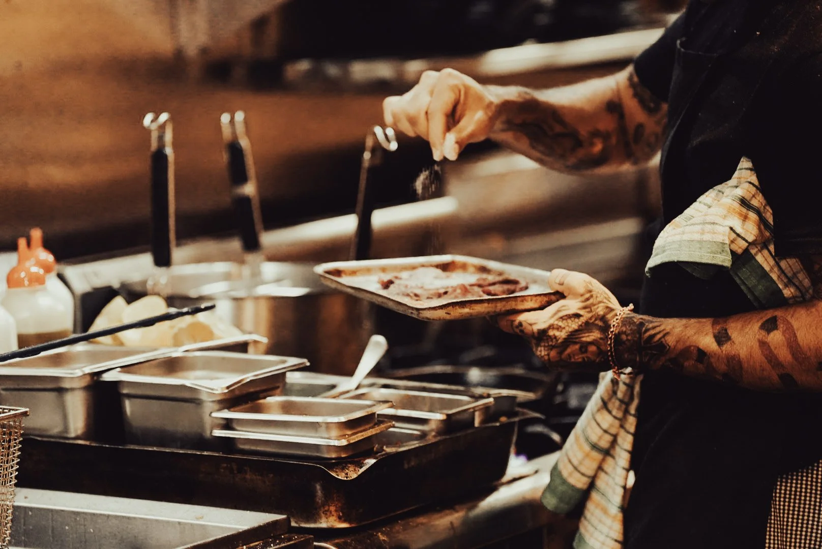 A person with tattooed arms seasoning or adding spice to a tray of raw meat in a professional kitchen, surrounded by cooking utensils and condiments.