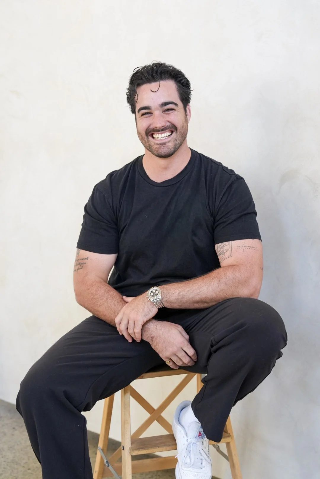 Smiling man with dark hair, beard, and tattoos, sitting on a wooden stool against a plain light-colored background, wearing a black t-shirt, black pants, white sneakers, and a silver watch.