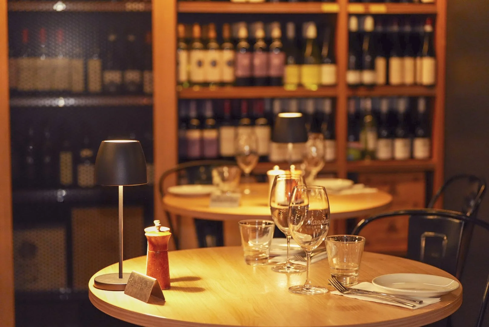 A cozy restaurant interior with a wooden table set for dining, including wine glasses, water glasses, plates, utensils, and a small red pepper shaker, illuminated by warm lighting from a table lamp, with shelves filled with bottles in the background.