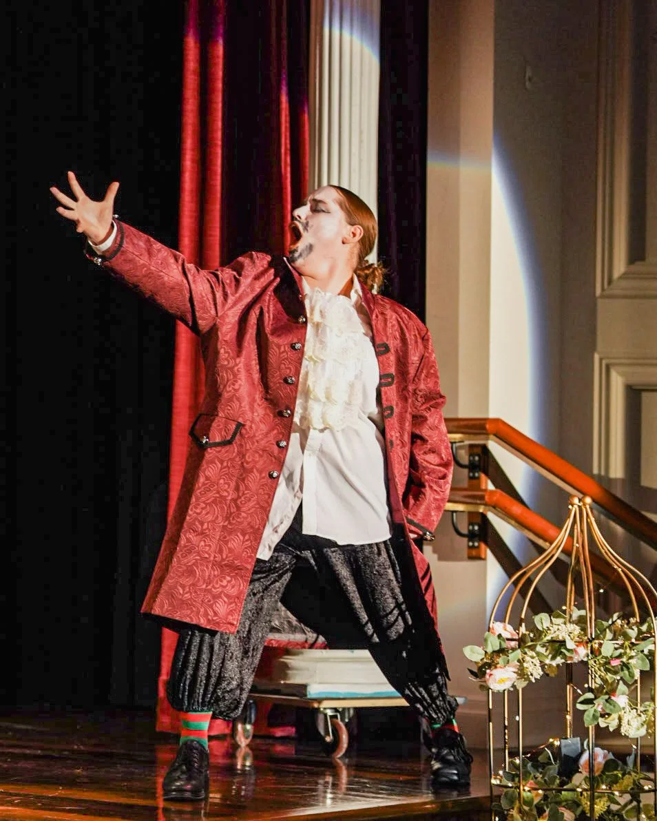 Performer on stage in period costume, wearing a red coat, white ruffled shirt, black textured pants, and colorful striped socks, striking a dramatic pose with one arm extended.