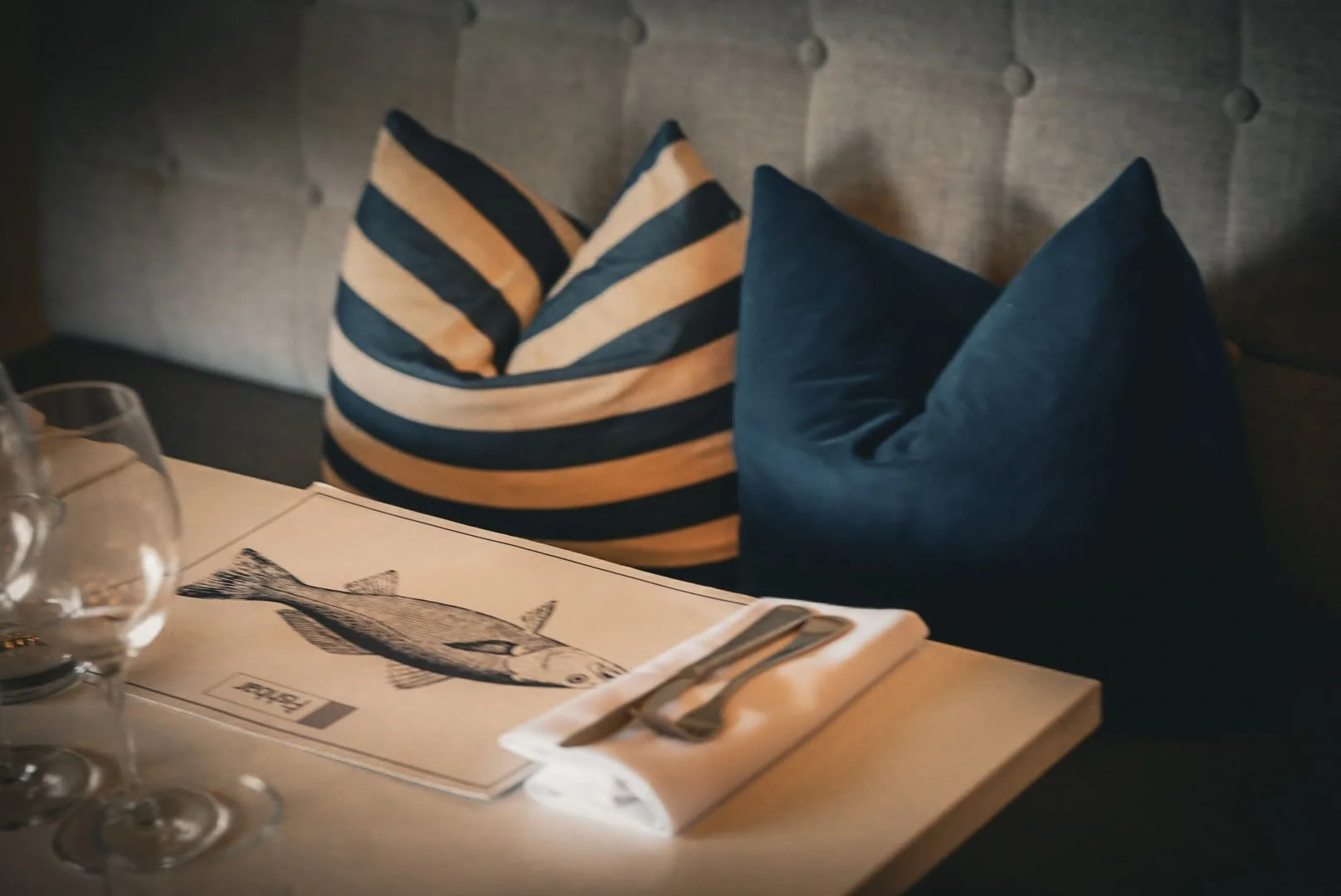 A restaurant table with drinking glasses and a menu featuring a fish illustration, set with silverware and napkins. There are decorative cushions on a gray upholstered bench in the background.