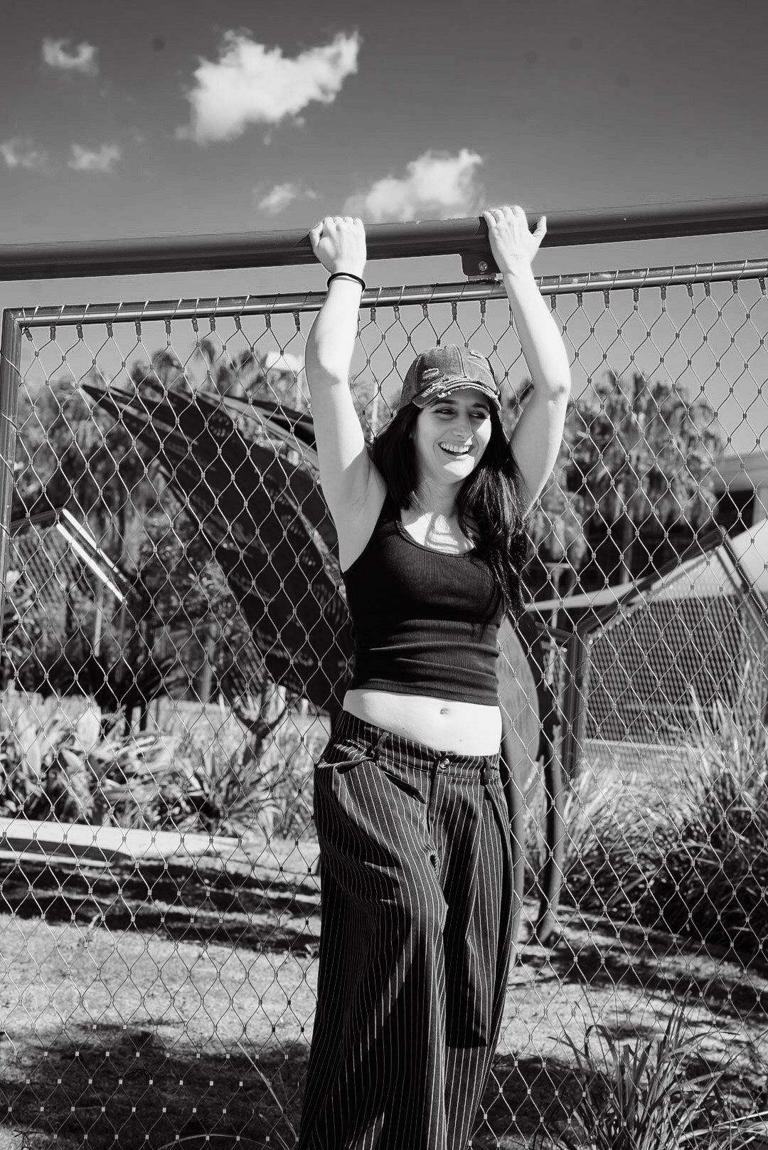 A young woman smiling and holding onto a fence outdoors on a sunny day, wearing a tank top, striped pants, and a cap.