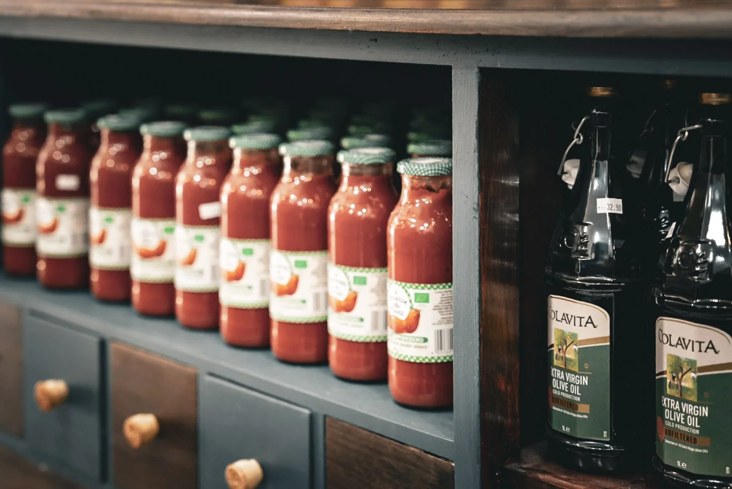 Shelf with multiple bottles of tomato sauce next to bottles of olive oil in a kitchen or grocery store.