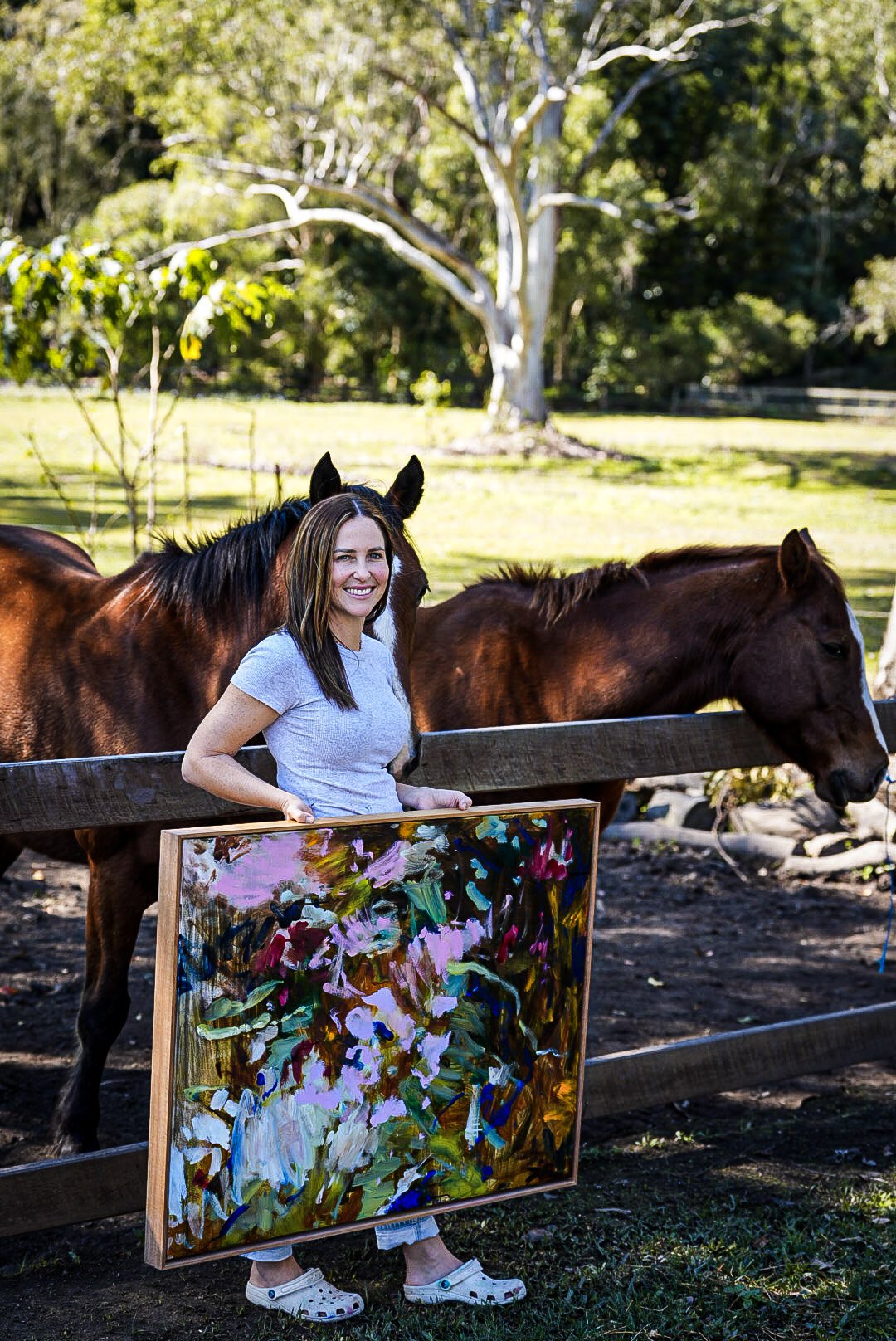 A woman holding an abstract floral painting standing outdoors beside two brown horses, with a green park and trees in the background.
