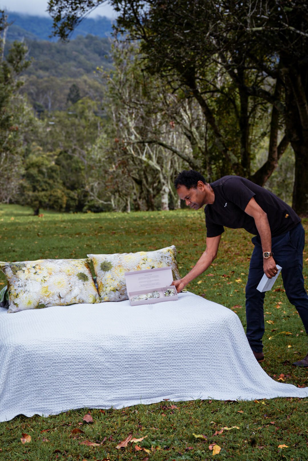 A man in black shirt and dark pants setting up a floral pillow and floral box on a white bedspread outdoors in a park with trees and mountains in the background.