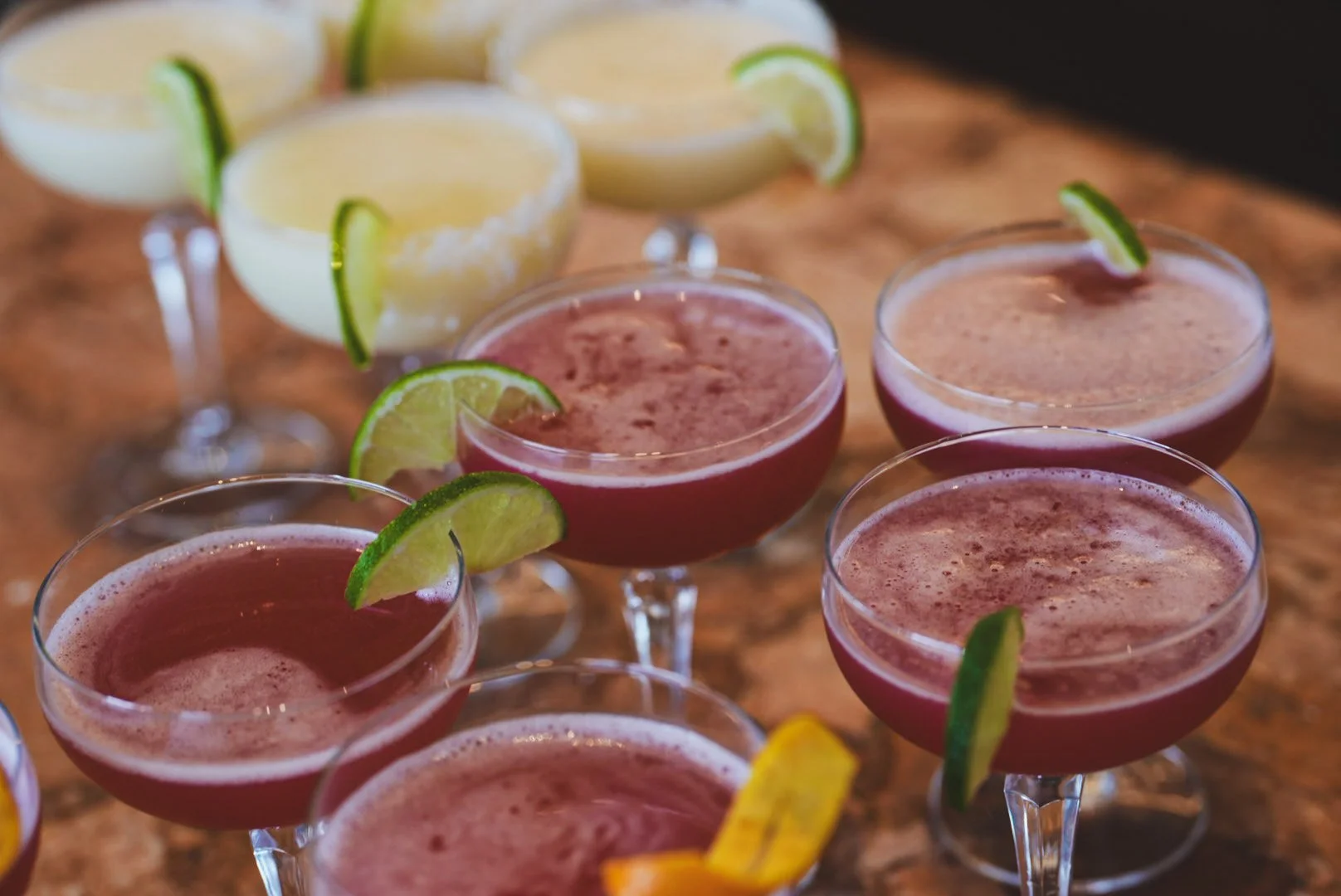 Cocktail glasses filled with pink and yellow drinks, garnished with lime and lemon slices, arranged on a wooden surface.