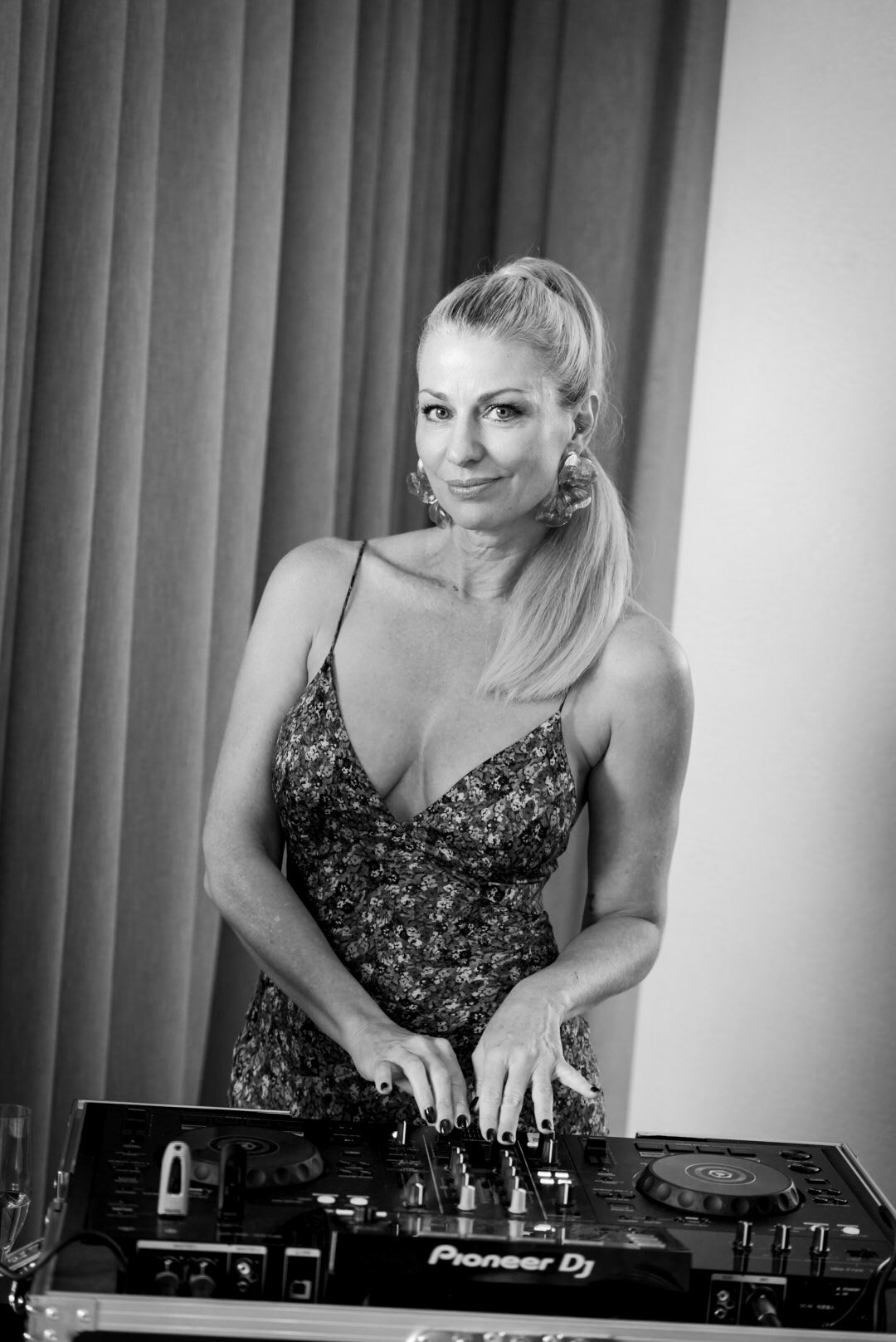 Black and white photo of a woman with blonde hair in a ponytail, wearing earrings and a floral dress, standing behind DJ equipment, smiling at the camera.