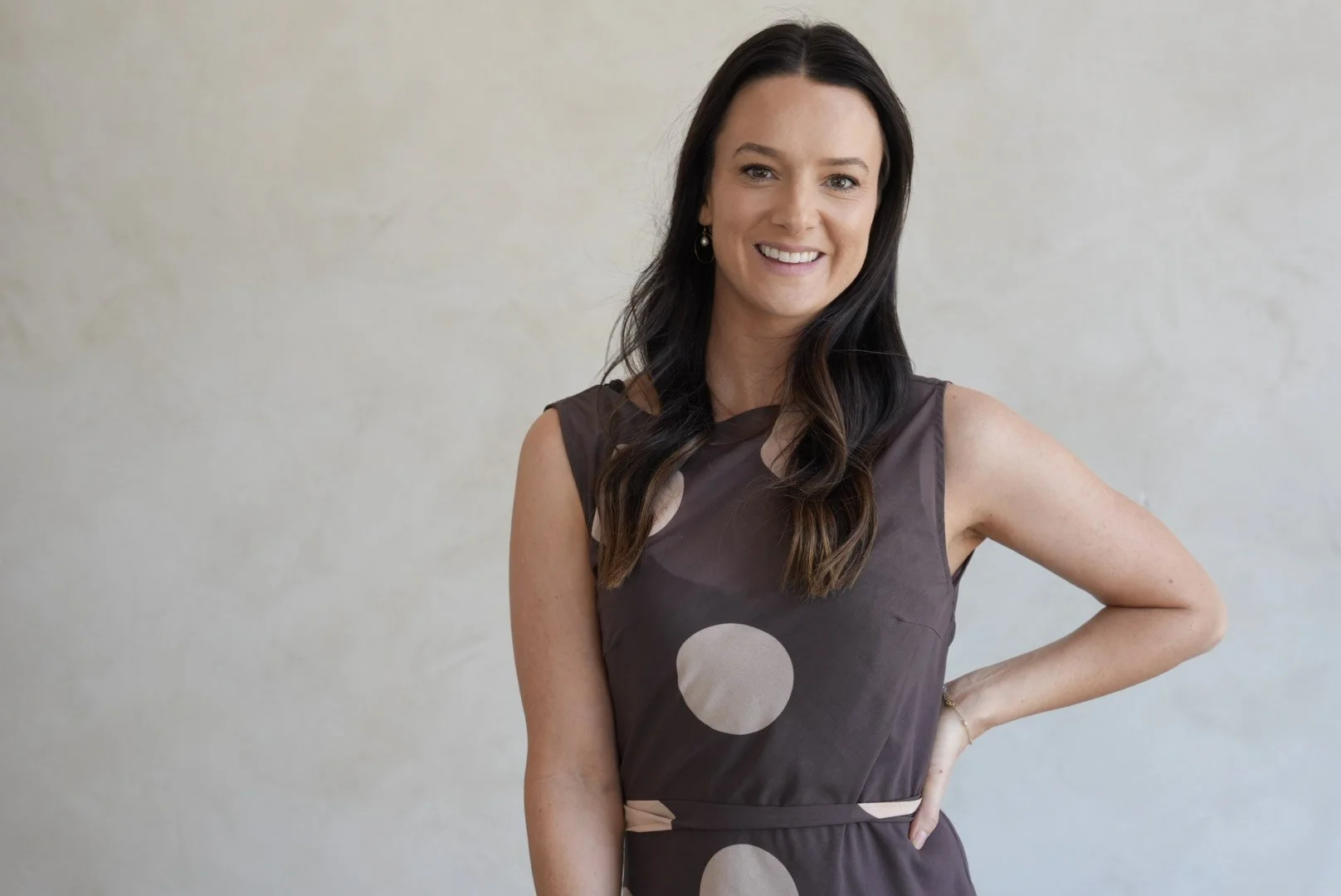 A woman with long dark hair wearing a sleeveless dress with large beige polka dots, smiling and standing against a plain light-colored background.