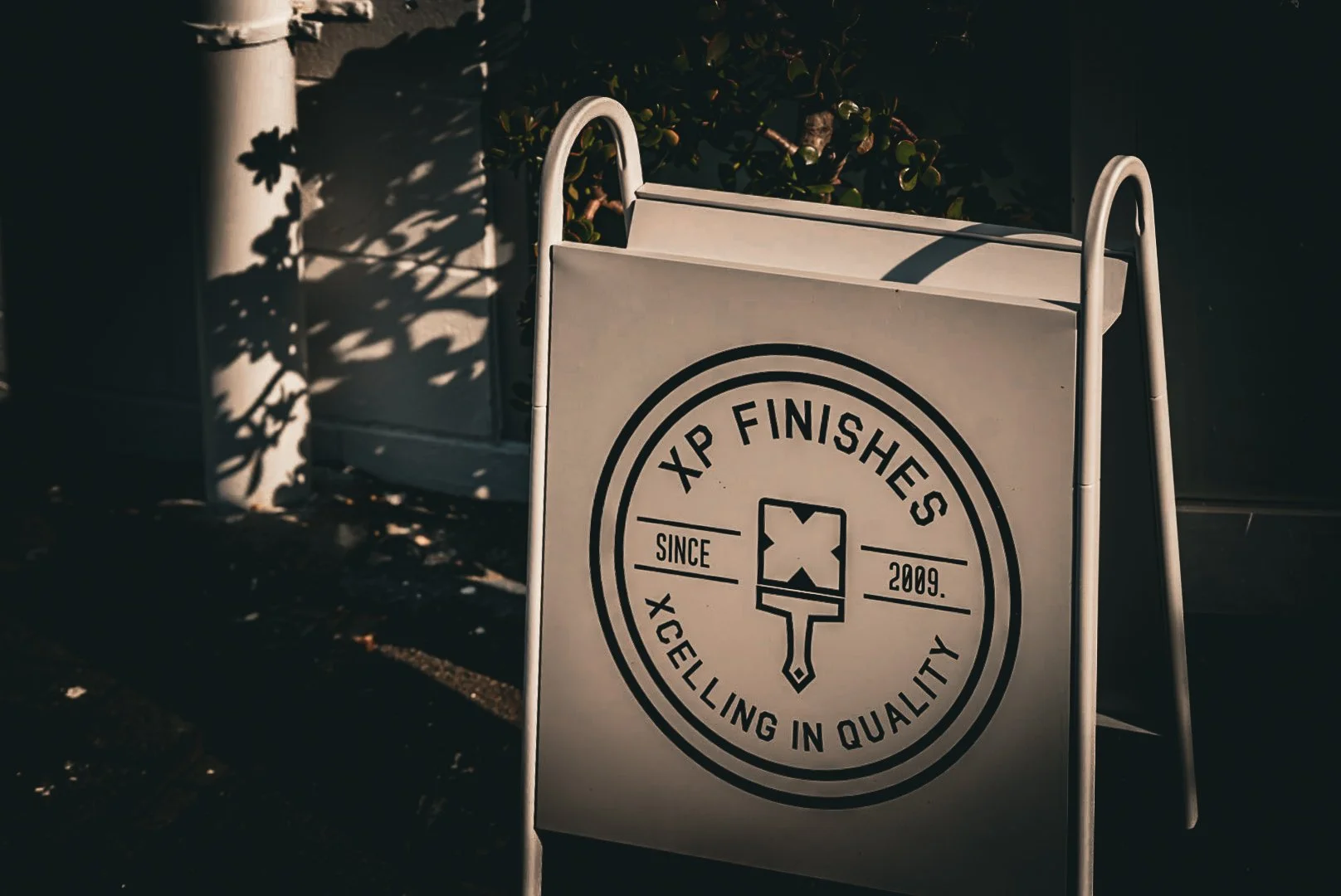 A white A-frame sidewalk sign with the text 'XP FINISHES SINCE 2009 XELLING IN QUALITY' and a popsicle illustration in the center. Shadows of leaves are cast on the sign and ground.