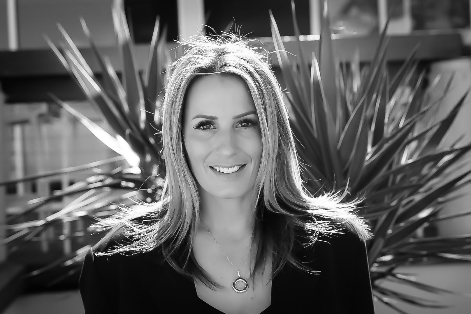 A smiling woman with long hair wearing a necklace against a backdrop of large plant leaves, in black and white.