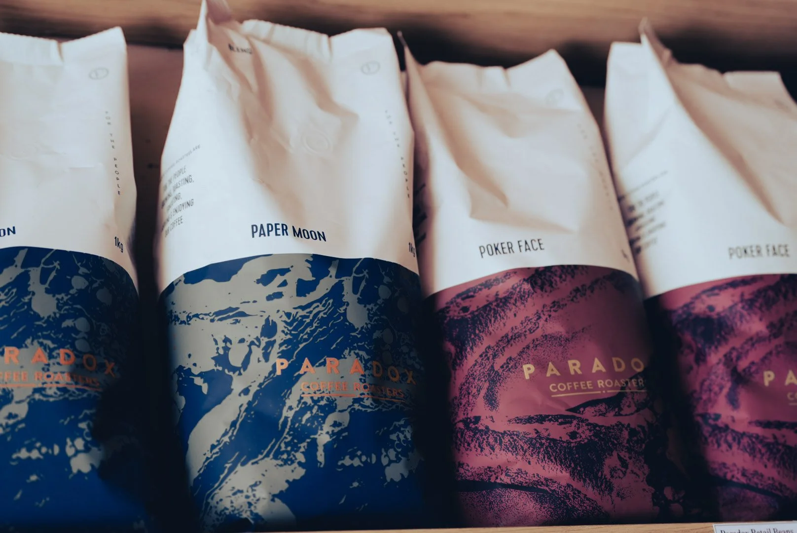 Four coffee bags lined up, two labeled 'Paper Moon' with a blue and white design, and two labeled 'Poker Face' with a pink and black design, all from Paradox Coffee Roasters.