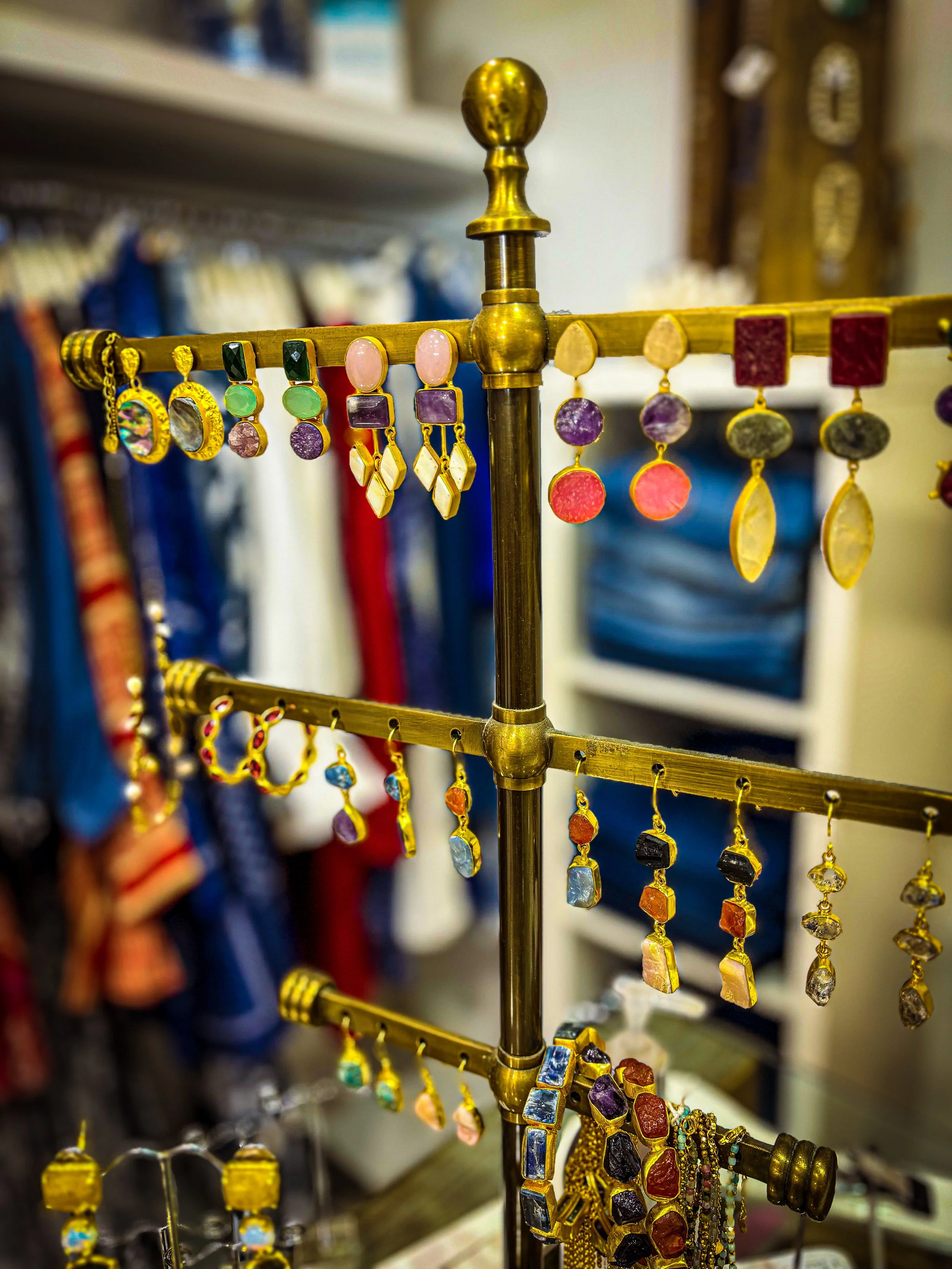 Gold jewelry display stand holding colorful earrings and necklaces, with blurred shelves of items in the background.