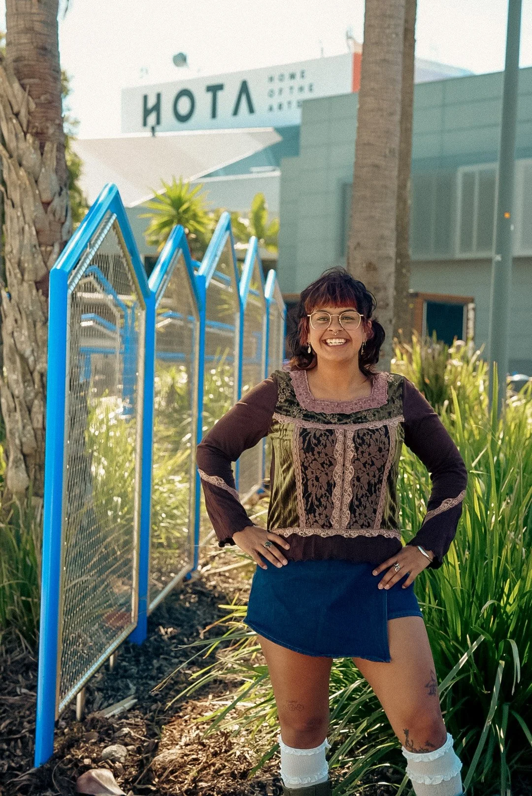 A woman with curly dark hair, wearing glasses, a floral embroidered top, a denim skirt, and white socks with dark shoes, standing outdoors with her hands on her hips, smiling at the camera. Behind her are green plants, a blue fence, palm trees, and a